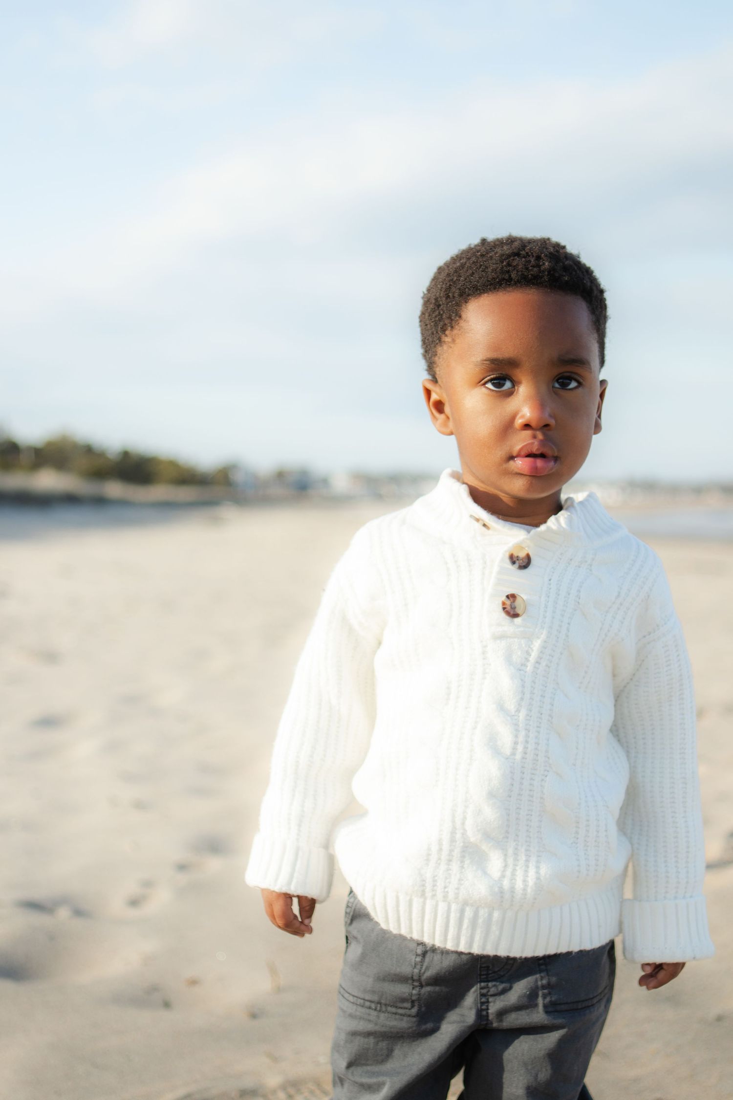 Young child wearing a white cable knit sweater stands on a beach during a sunny day.