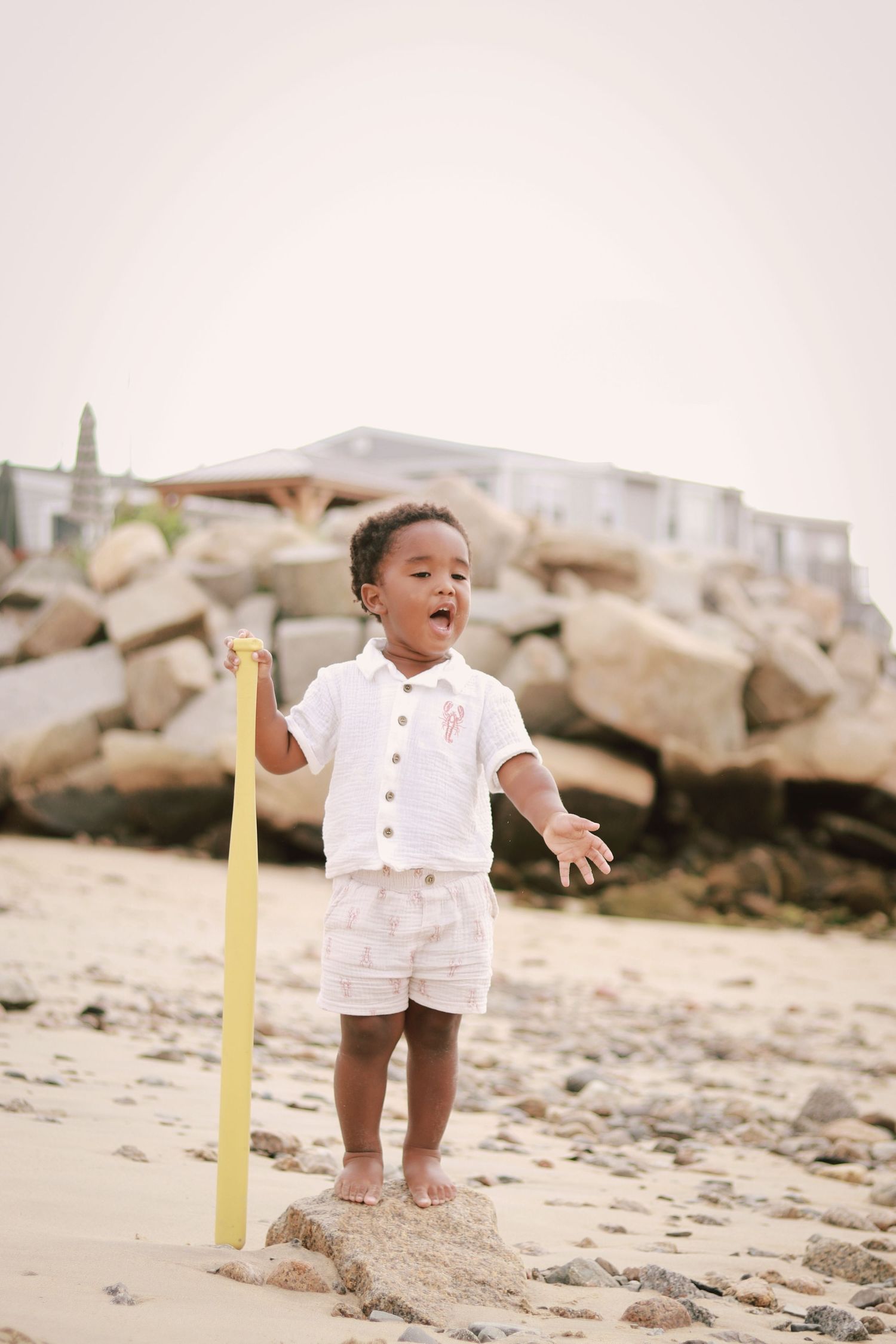 Young child in white outfit playing joyfully on a sandy beach near rocks.