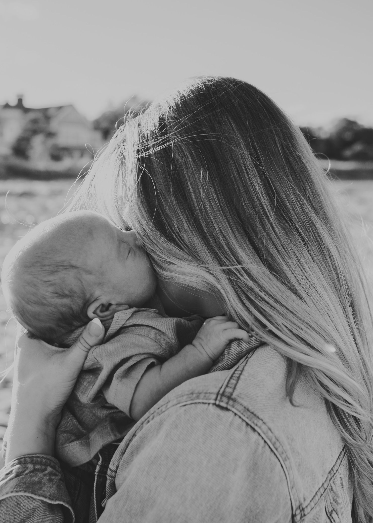 Black and white photo of a mother with long blonde hair tenderly holding and kissing her newborn baby outdoors.