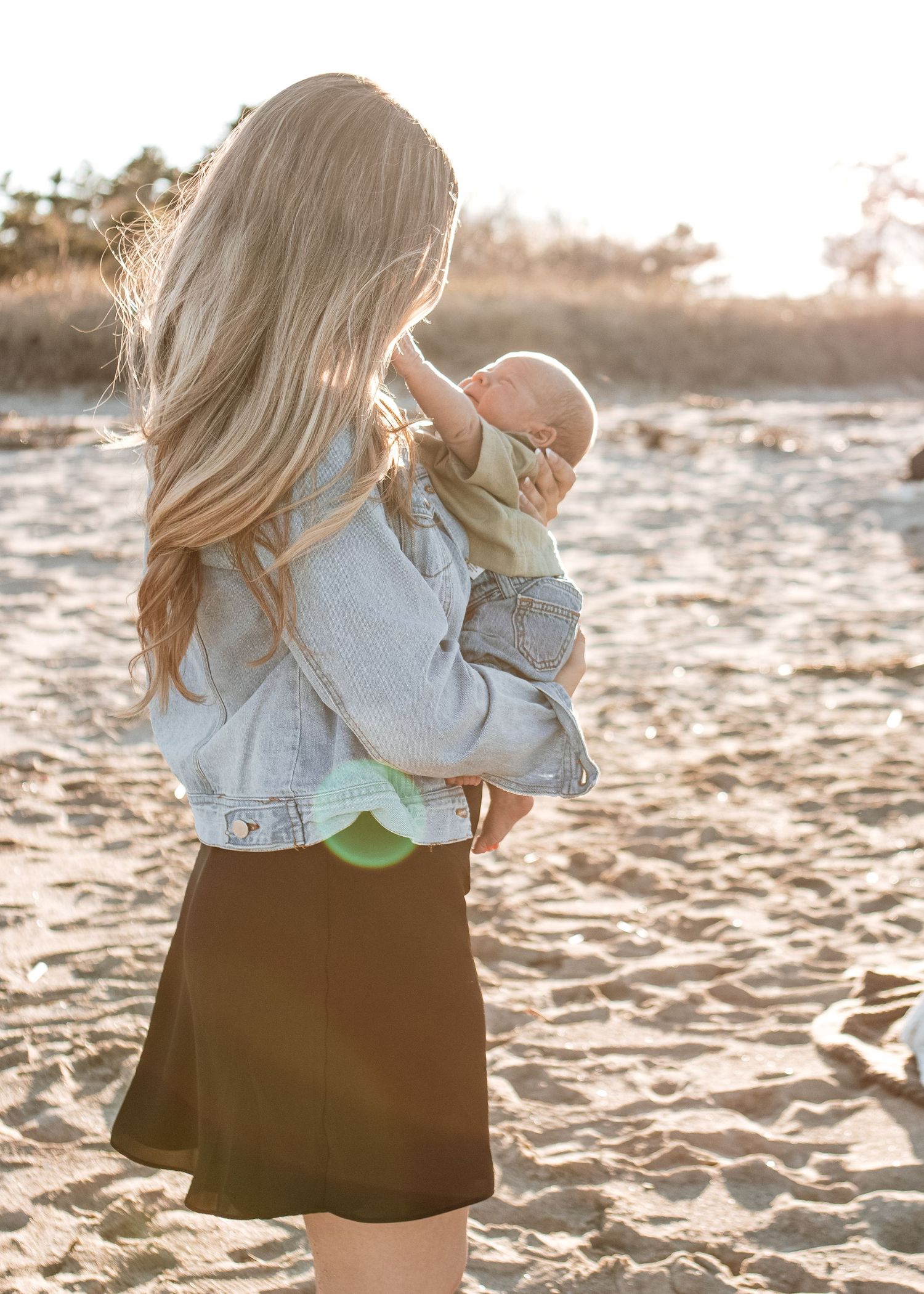 A blonde woman in a denim jacket holds a baby on a sandy beach in warm golden sunlight.