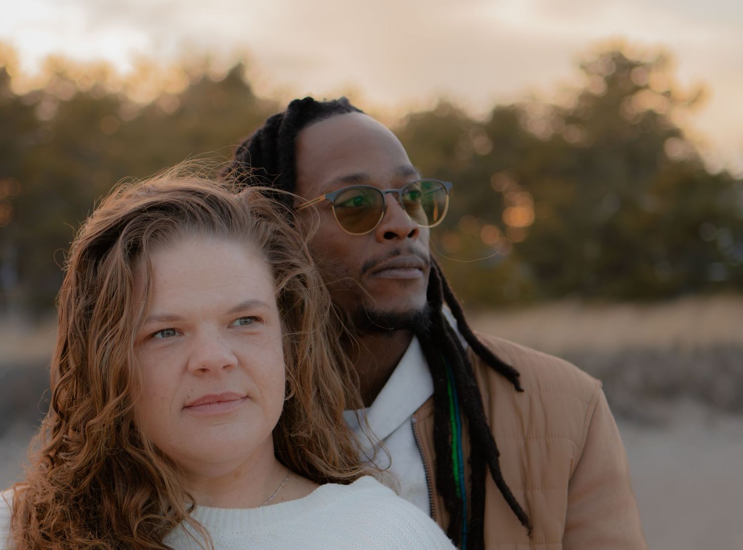 A couple poses together outdoors at sunset, woman with curly hair and man with dreadlocks and sunglasses.