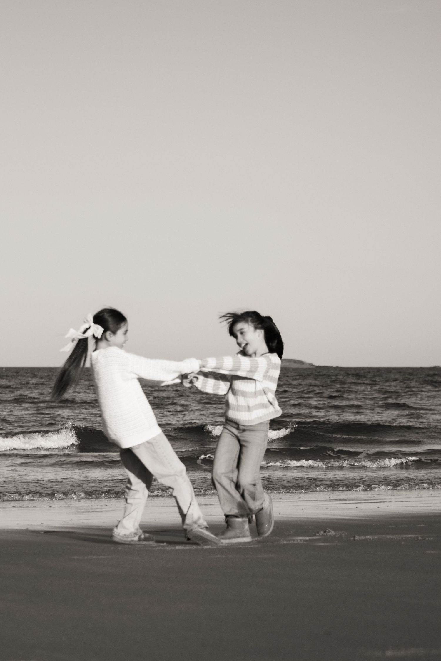 Two girls holding hands and spinning on a beach shore in black and white photo.