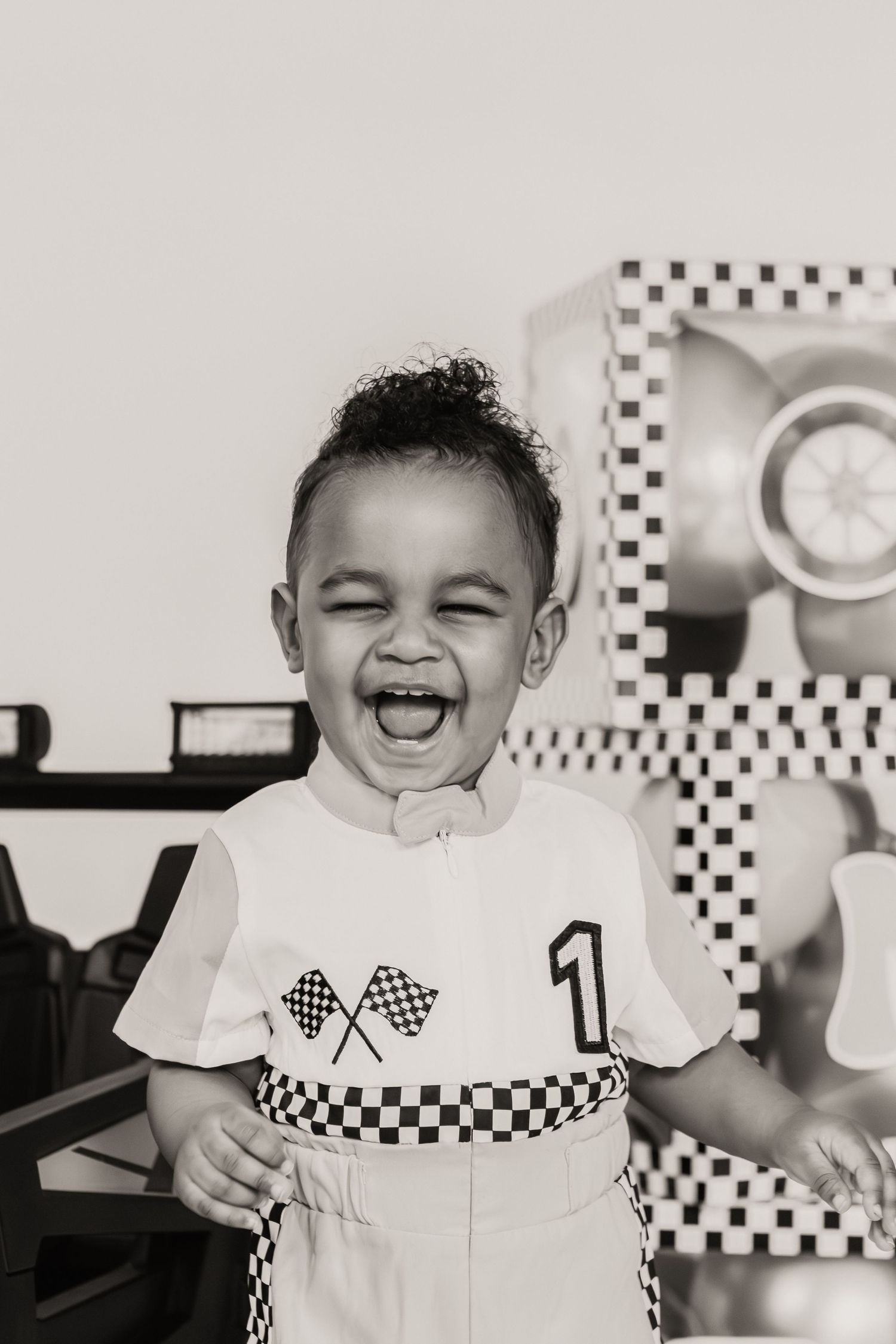 Black and white photo of a joyful toddler in a racing-themed outfit with checkered flags, laughing at a birthday party.