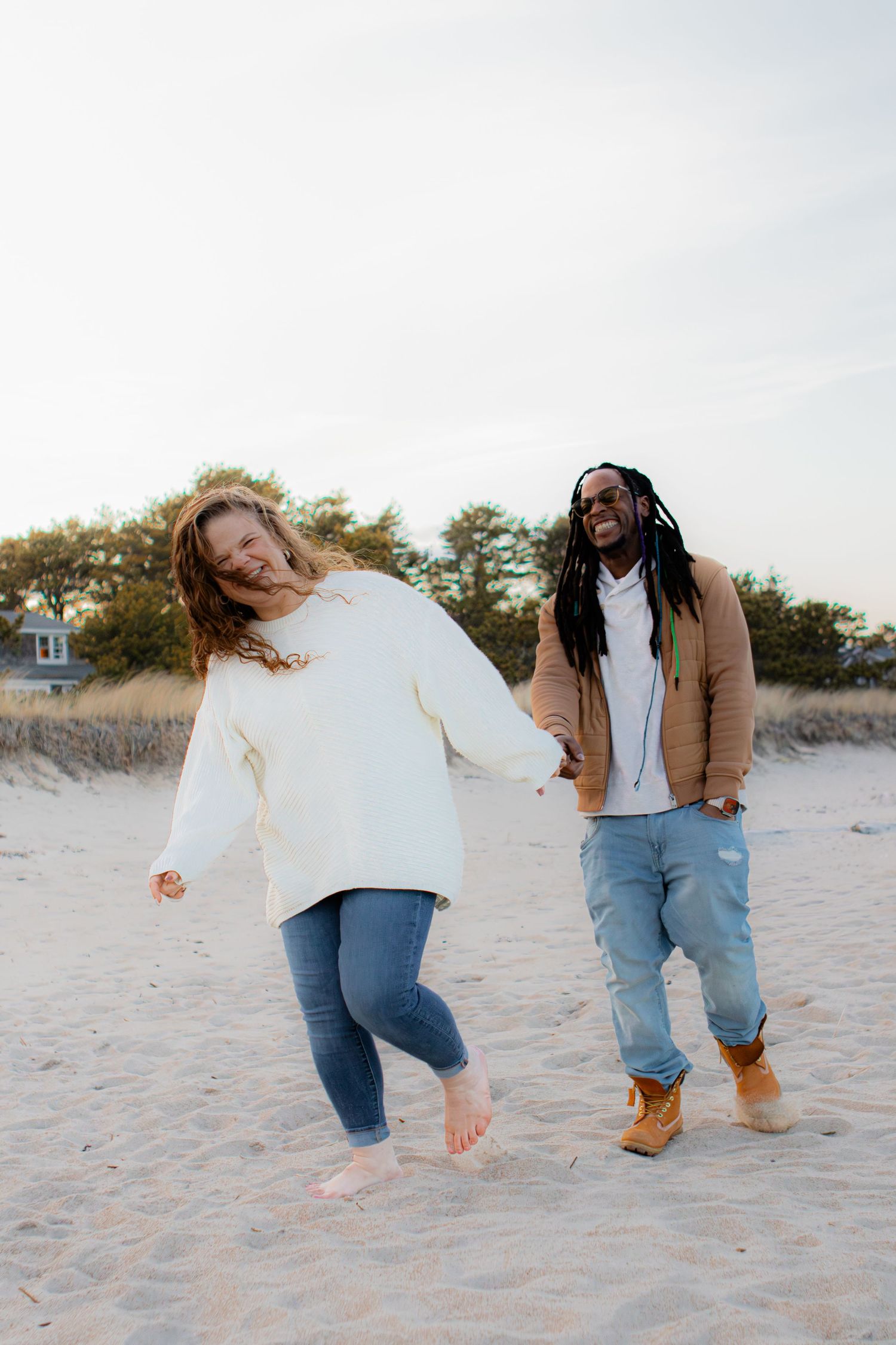 A joyful interracial couple holds hands and laughs while walking barefoot on a sandy beach in casual fall outfits.