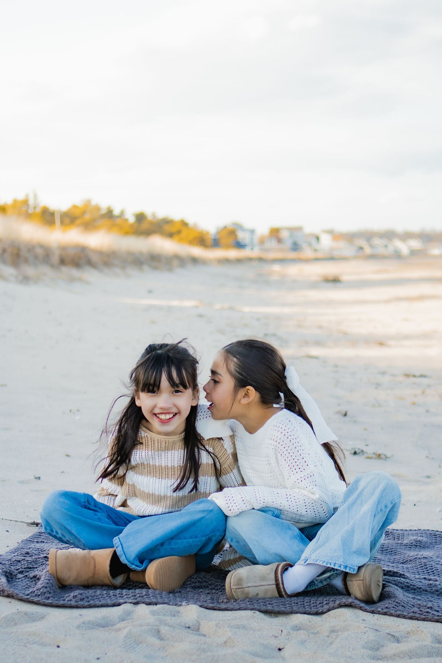Two girls sitting on a blanket at the beach, one whispering in the other's ear, both wearing jeans and sweaters.