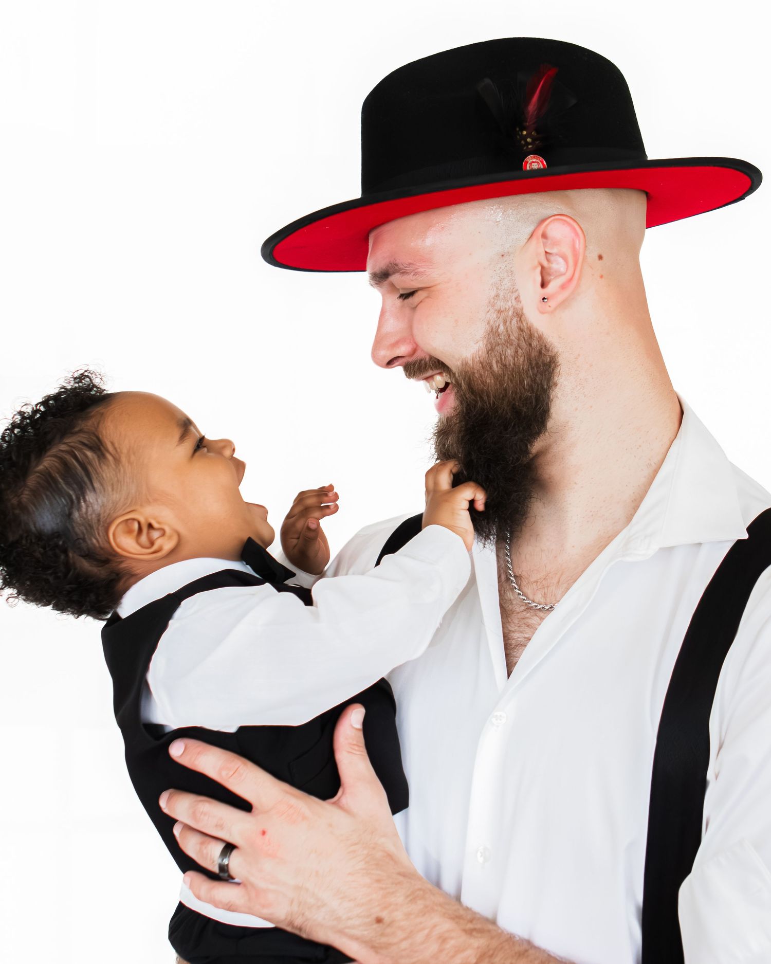 A smiling bearded man wearing a black and red wide-brim hat holds a laughing toddler in a white shirt and suspenders.