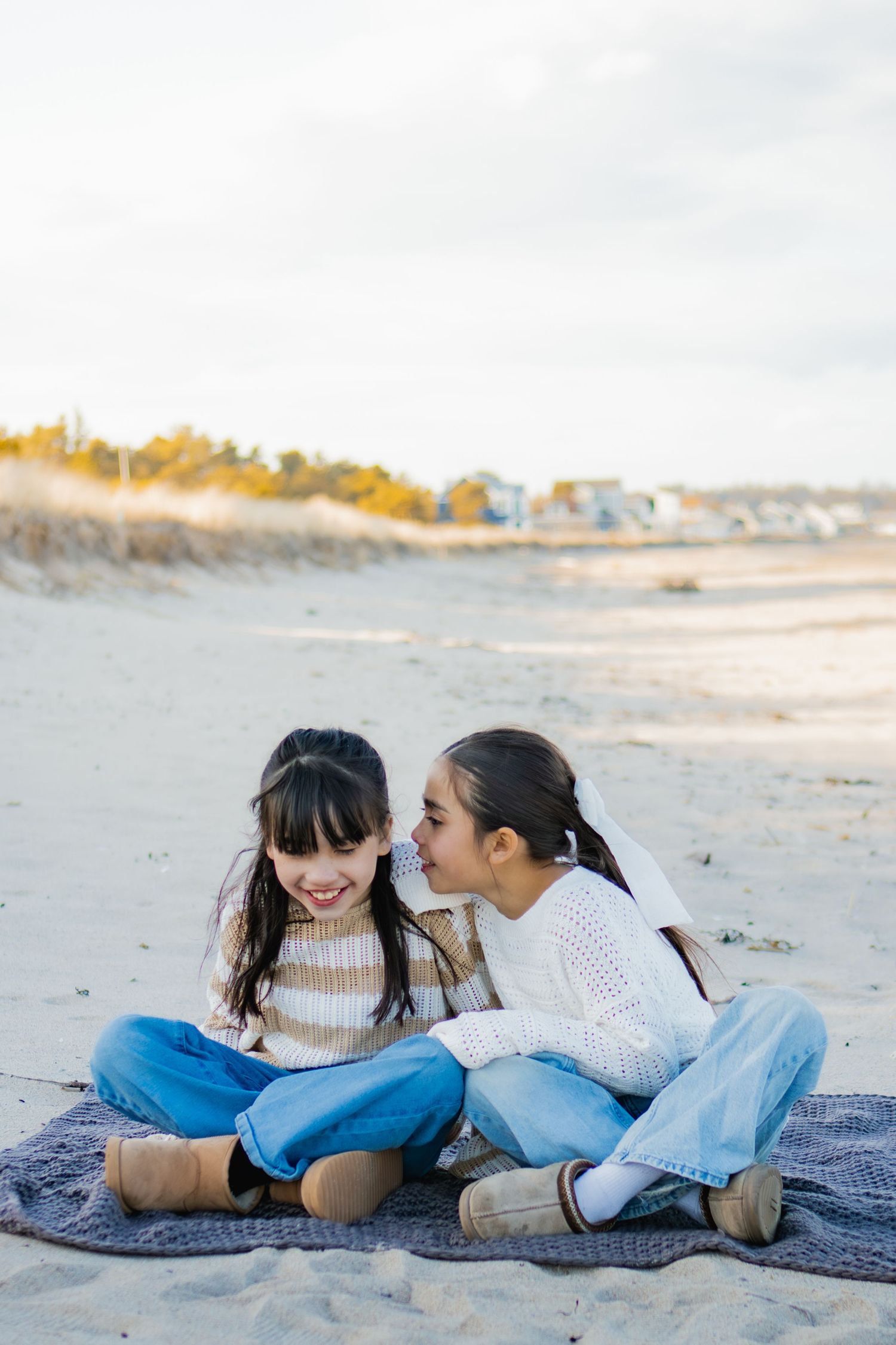 Two girls sitting on a blanket on the beach, sharing secrets and smiling, wearing cozy sweaters and jeans.