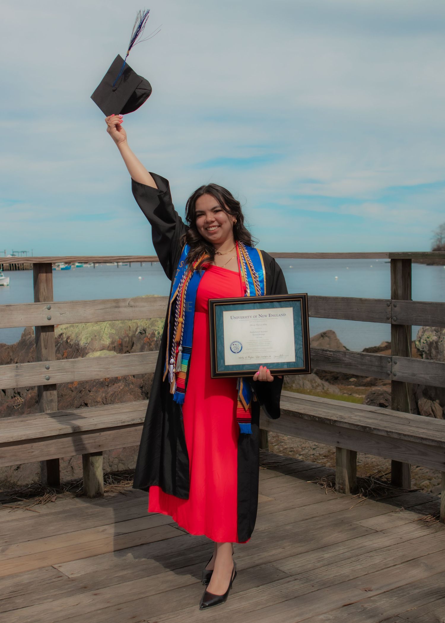 Woman in graduation gown and cap celebrating with diploma by waterfront boardwalk wearing red dress.