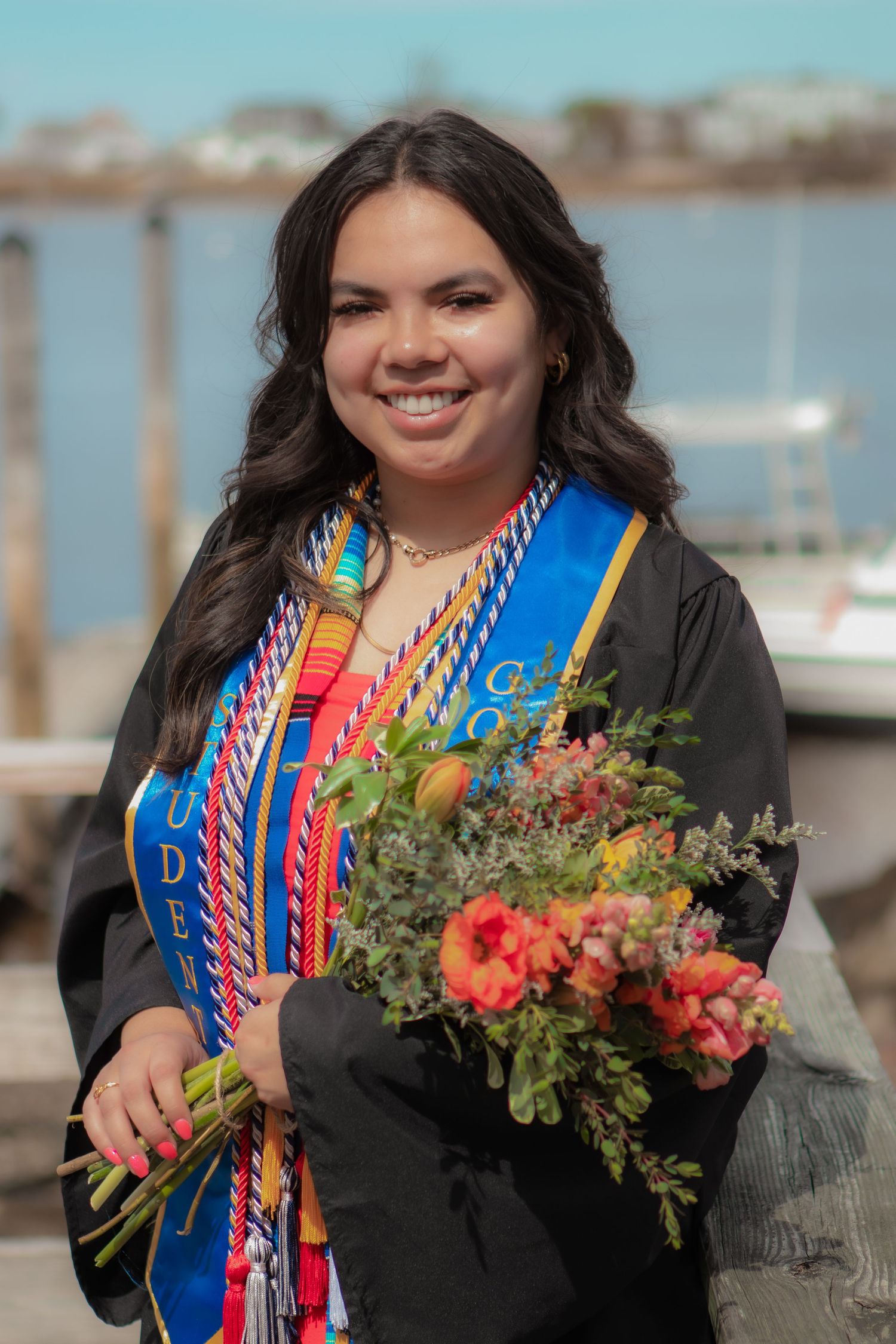 Smiling graduate in black gown with colorful sashes holds floral bouquet near waterfront on graduation day.