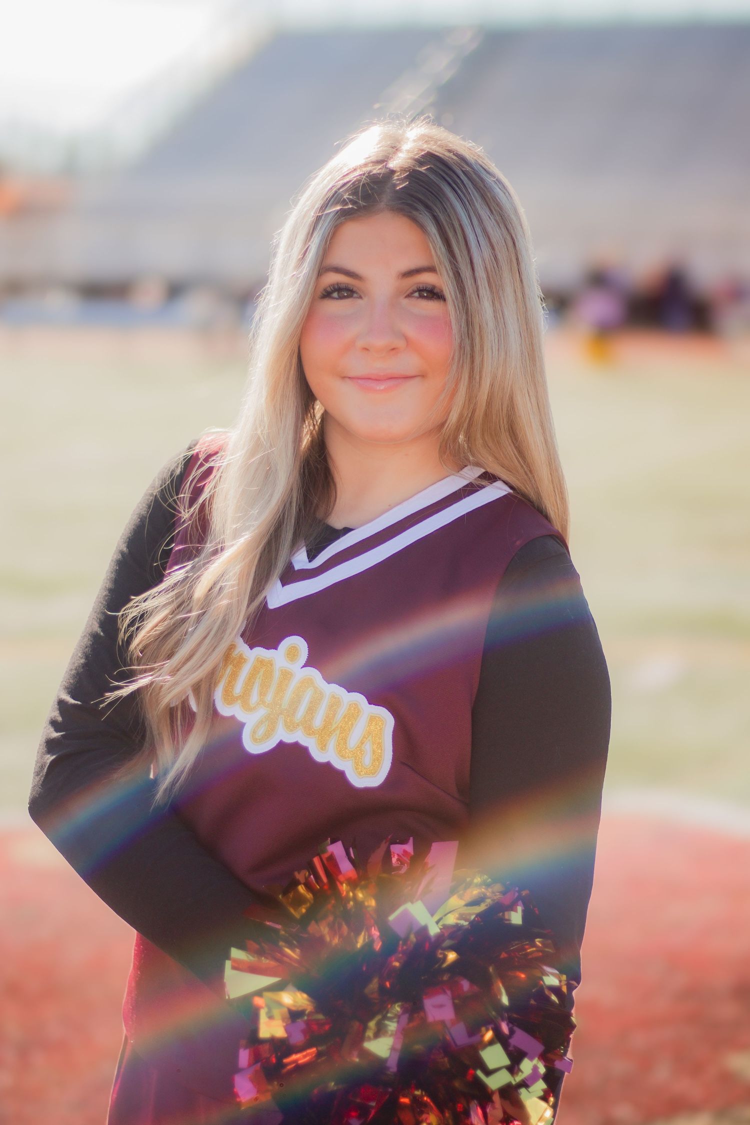 Cheerleader in maroon uniform poses with pom poms in sunlit outdoor setting.