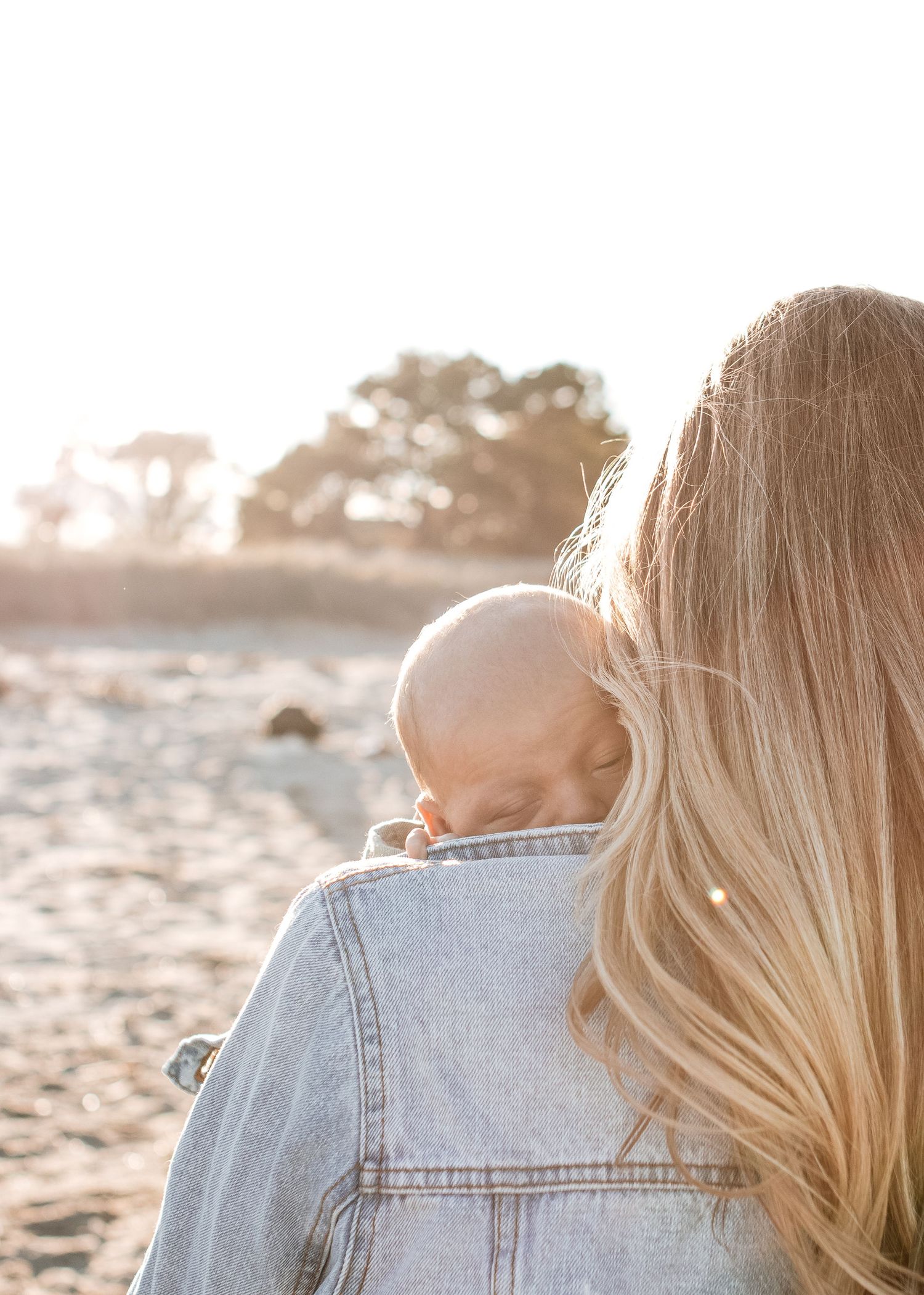 Mother in denim jacket holding sleeping baby on sunlit beach.