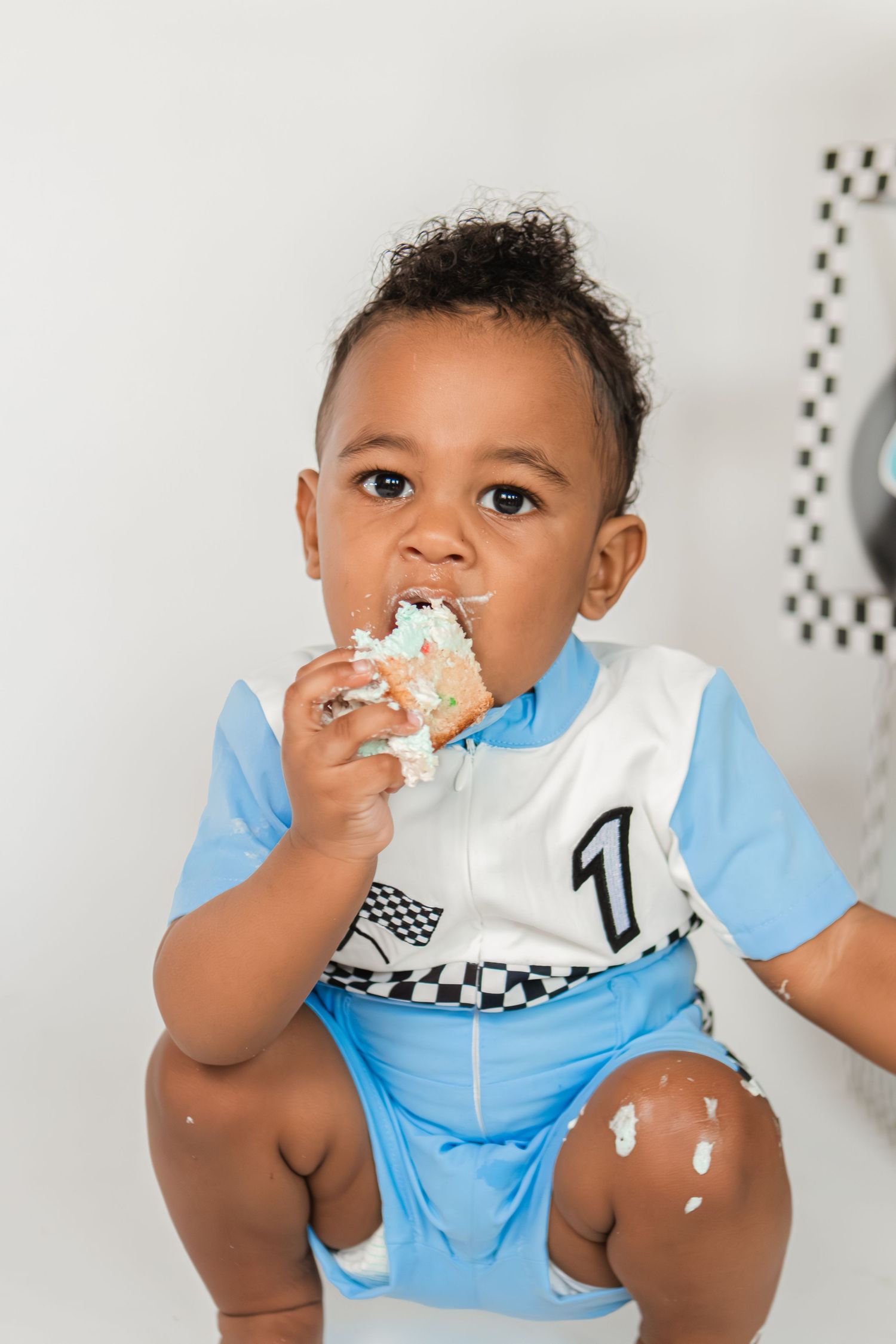 Adorable baby boy eating smash cake in blue racing outfit during first birthday photo shoot.