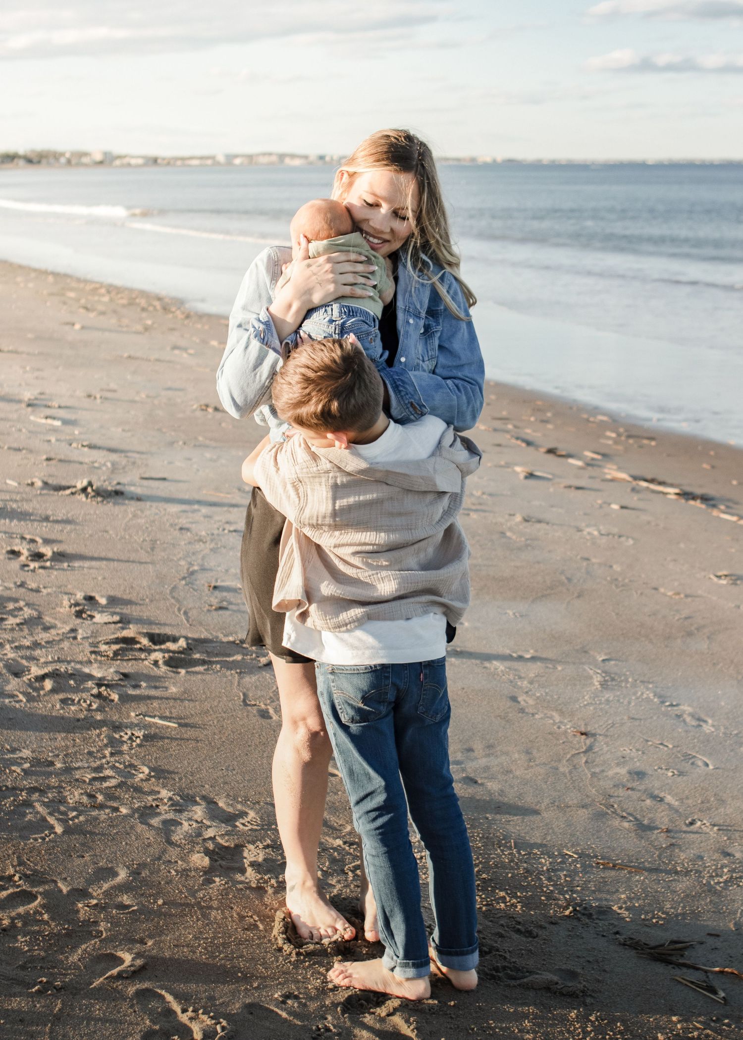 A family of three shares a tender moment on a sandy beach, with a mother holding a newborn while a child embraces her.