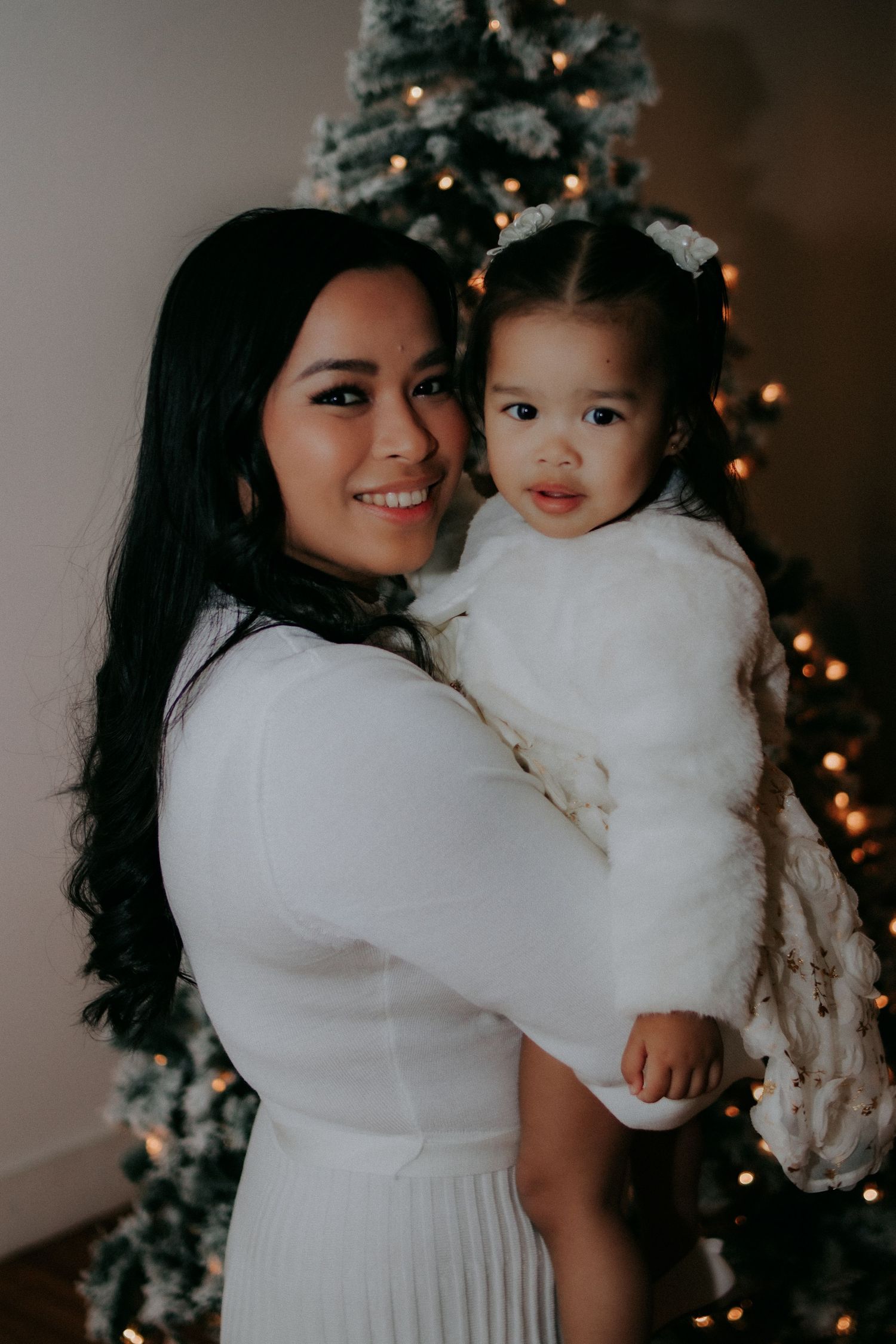 Mother and daughter share tender holiday moments in white dresses against a Christmas tree backdrop.