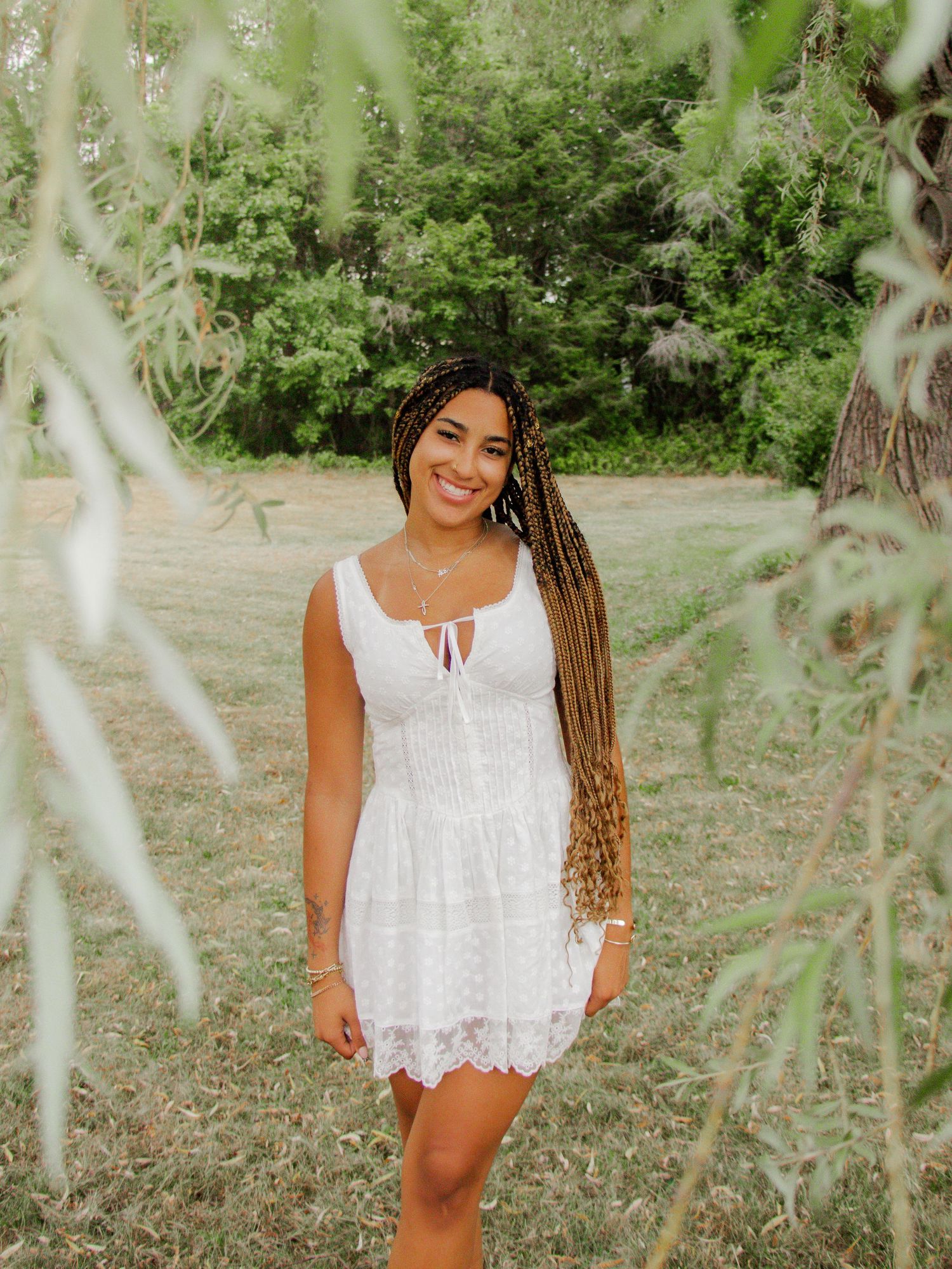 Woman in white lace top smiling outdoors among green foliage.