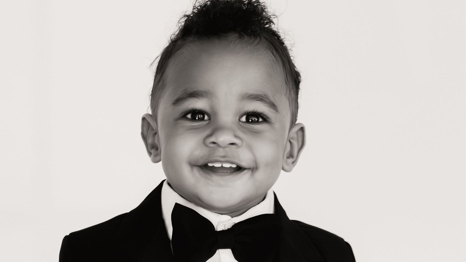 Adorable toddler boy smiling in a black tuxedo with bow tie, black and white portrait photo.