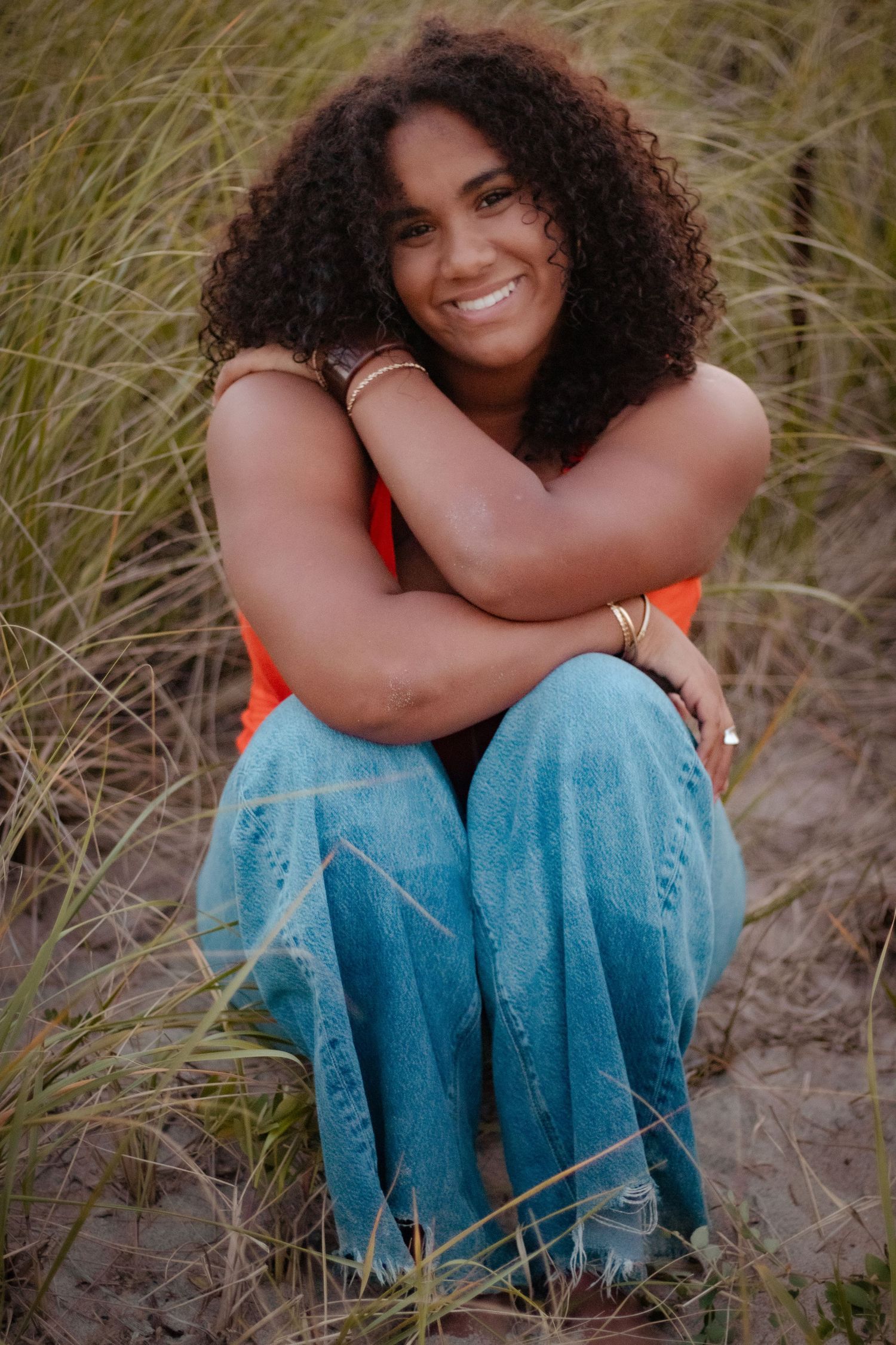 Person in orange top and jeans sits among beach grass during outdoor photoshoot.
