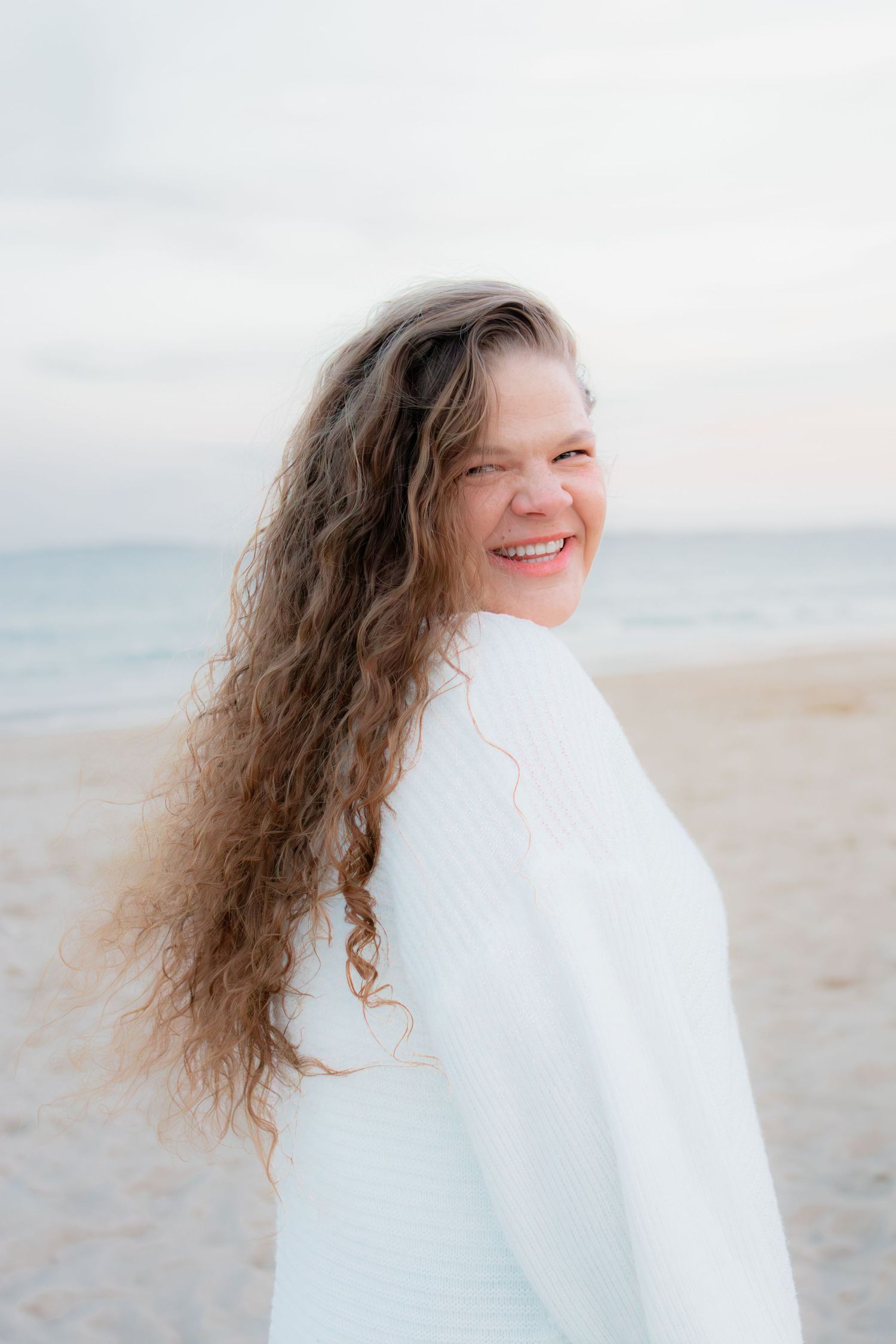 Smiling woman with long curly hair wearing white sweater on a beach at sunset.