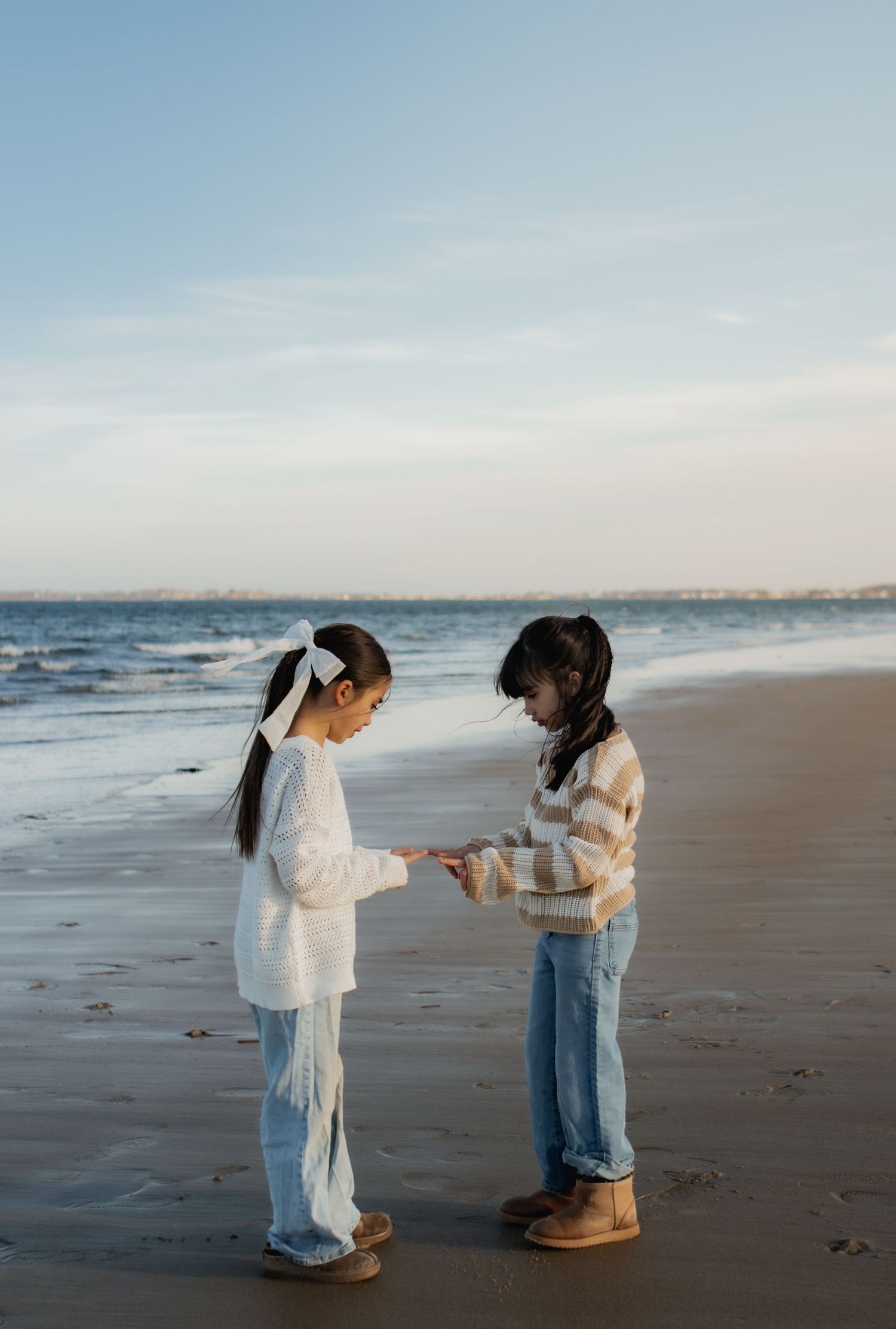 Two young girls in jeans and cozy sweaters examine a small object together on a sandy beach by the ocean.