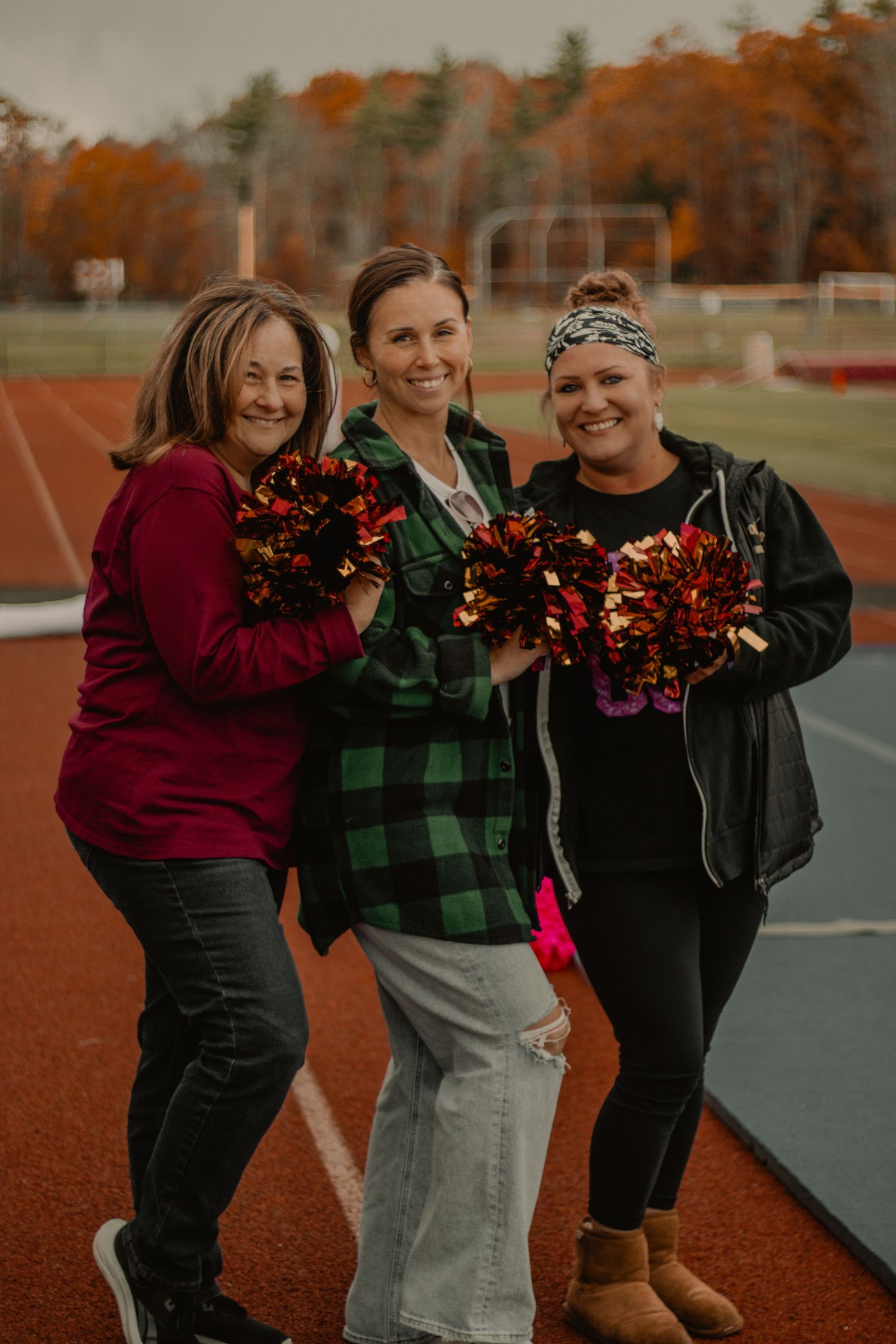 Three cheerleaders holding pom poms pose together on a track field during autumn.