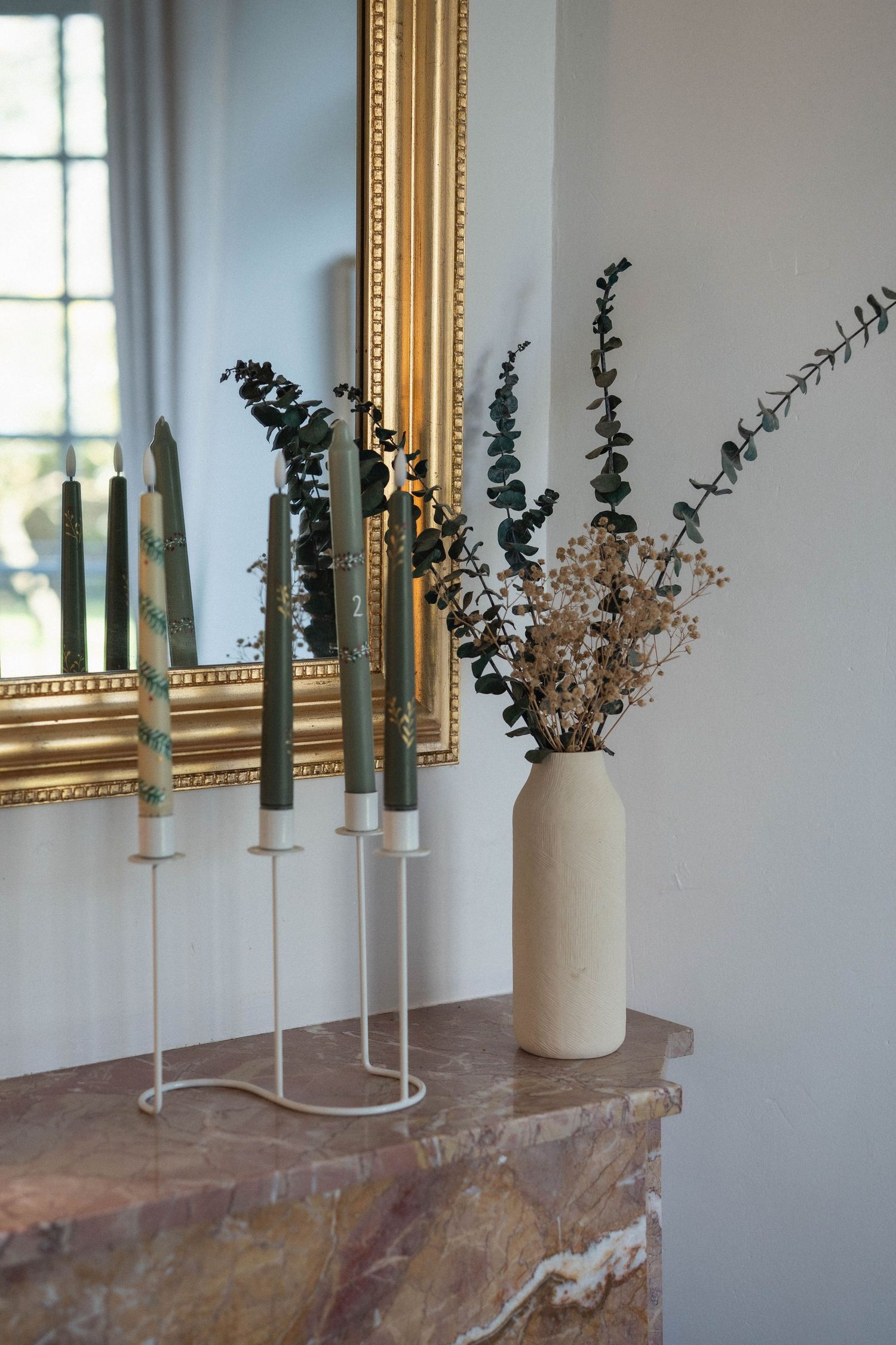 Modern home decor featuring green candles and dried flowers in a white vase on a marble surface.