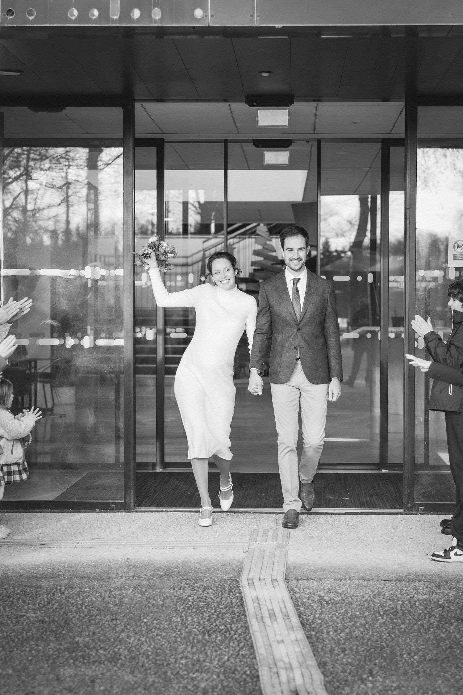 Black and white photo of a couple exiting a modern building together.
Sortie de la mairie des jeunes mariés en noir et blanc.