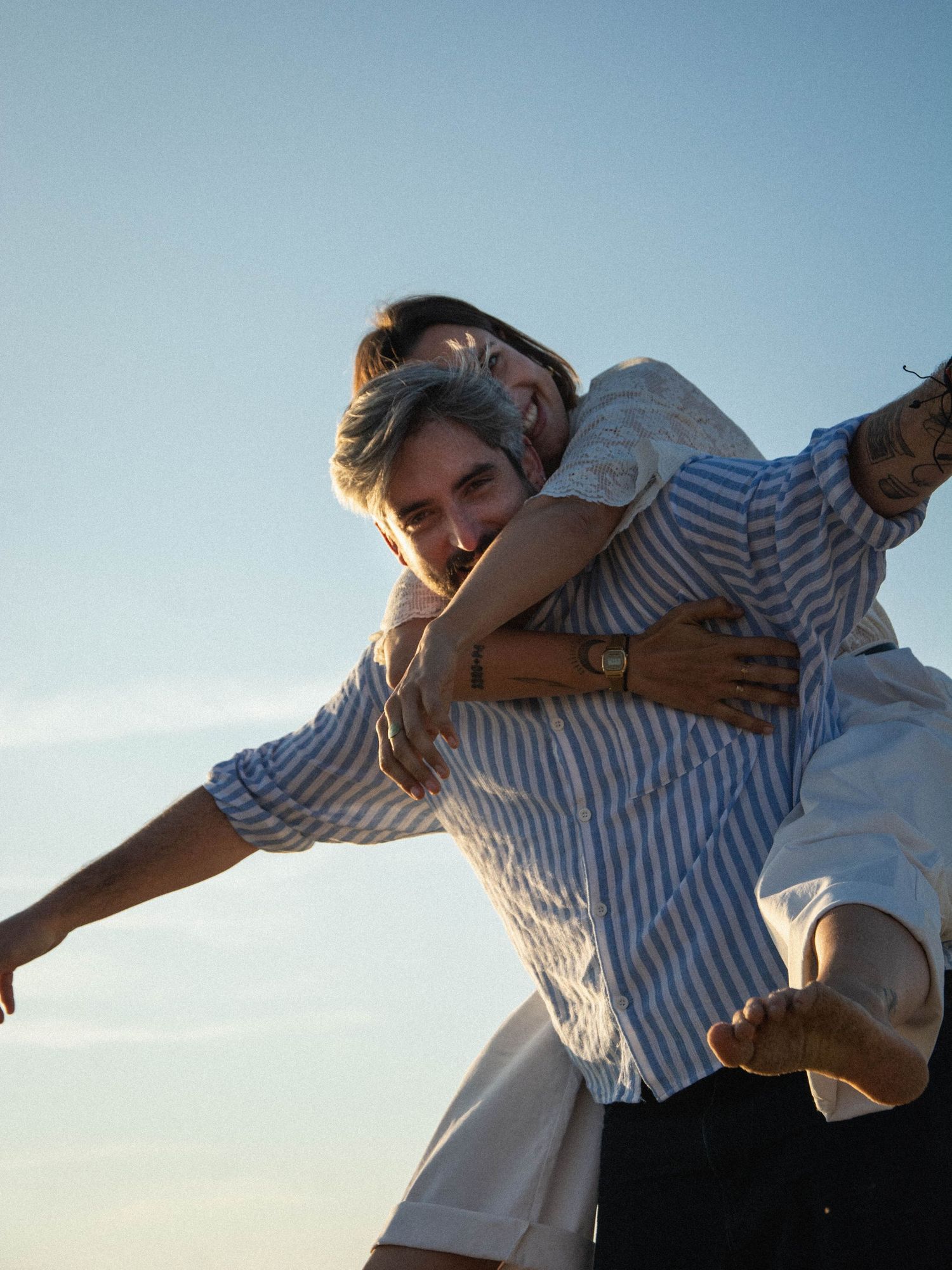 A playful couple dancing and embracing against a bright blue sky.
Couple souriant avec femme sur le dos et bras en avion de l'homme.