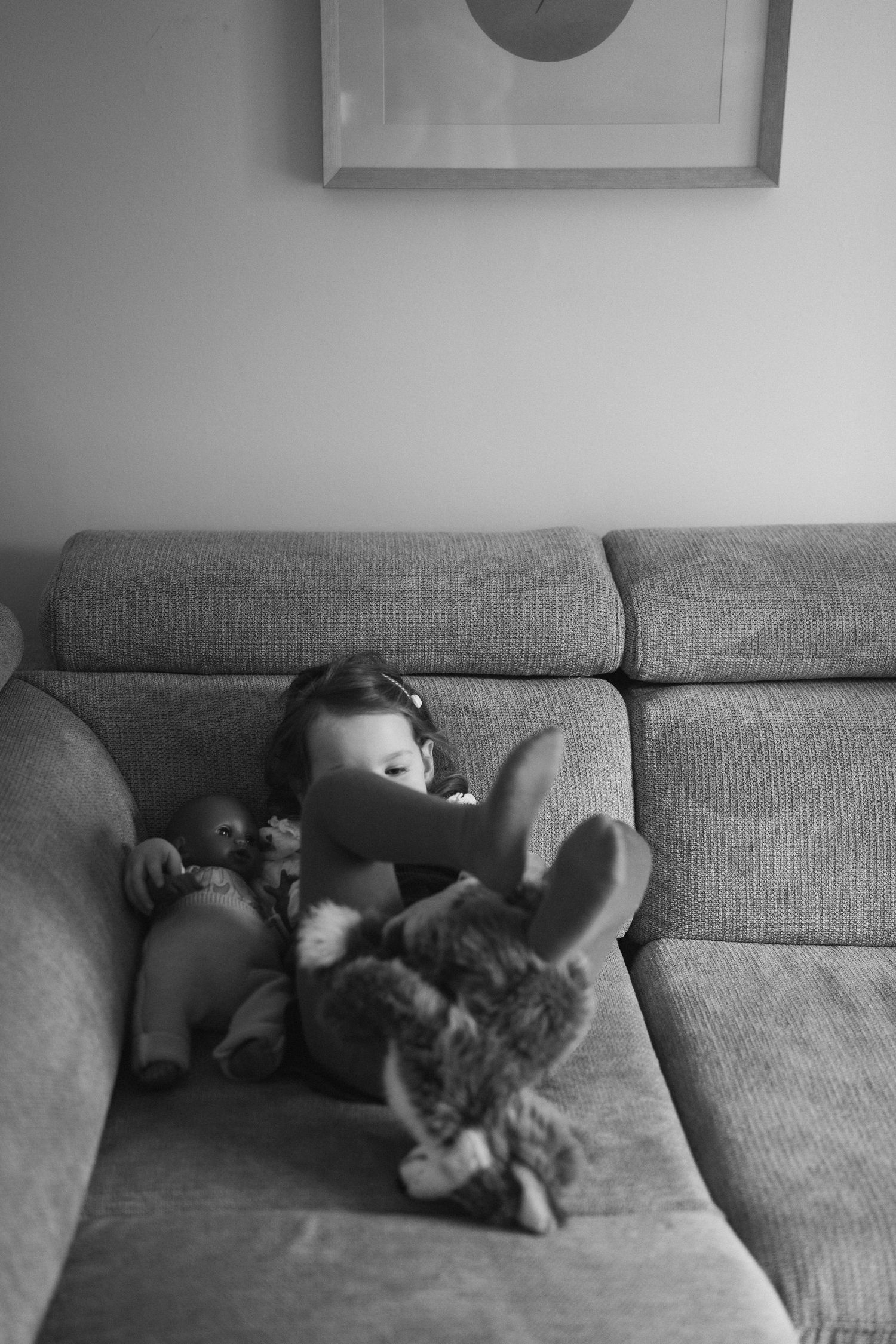 Photo noir et blanc d'une petite fille sur le canapé avec son doudou et sa poupée.