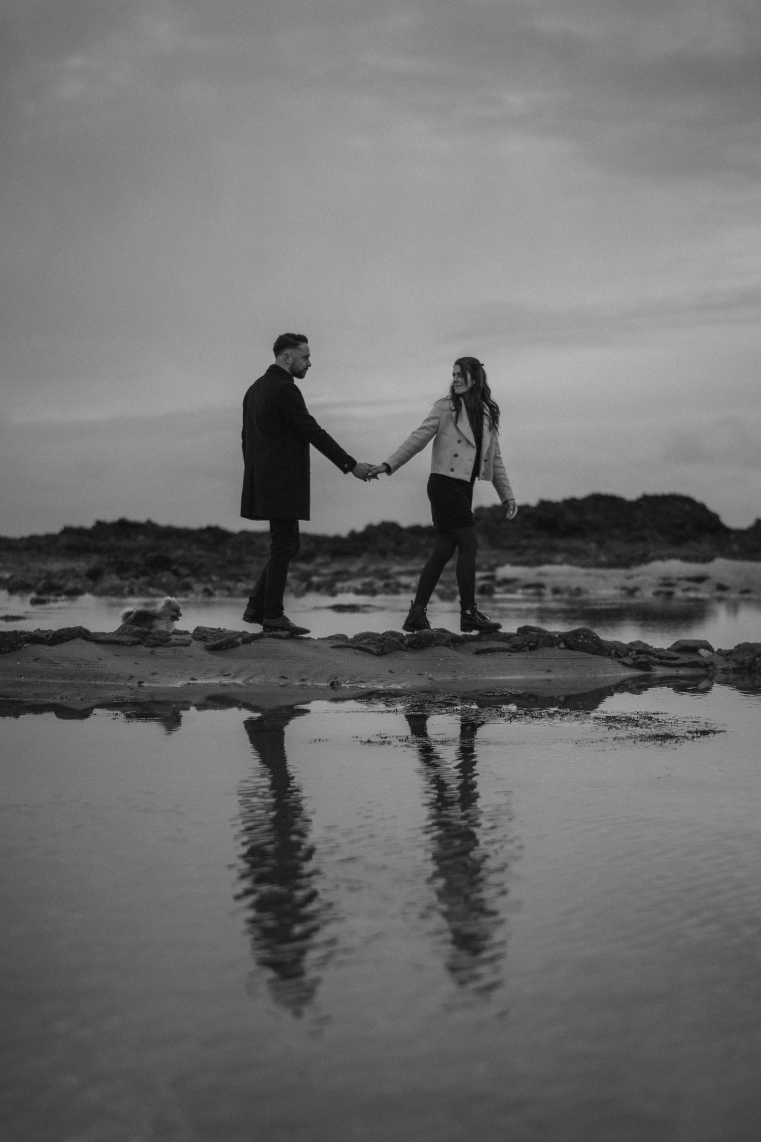 Black and white photo of couple holding hands on rocky beach with reflection in water.
Couple se tenant la main et leur reflet.
