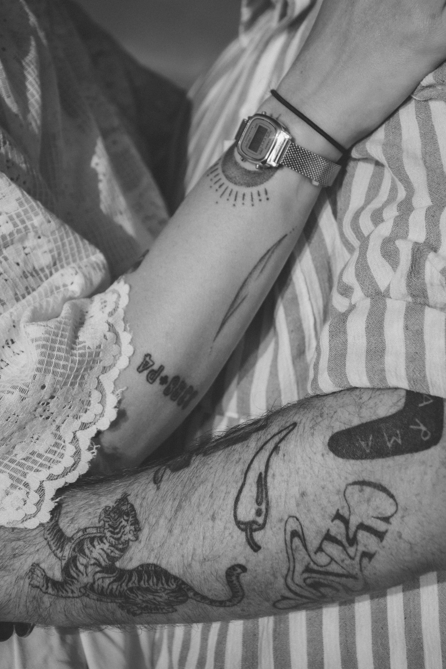 Black and white close-up of tattooed arms with intricate designs on striped fabric.
Photo en noir et blanc des bras tatoués d'un couple.