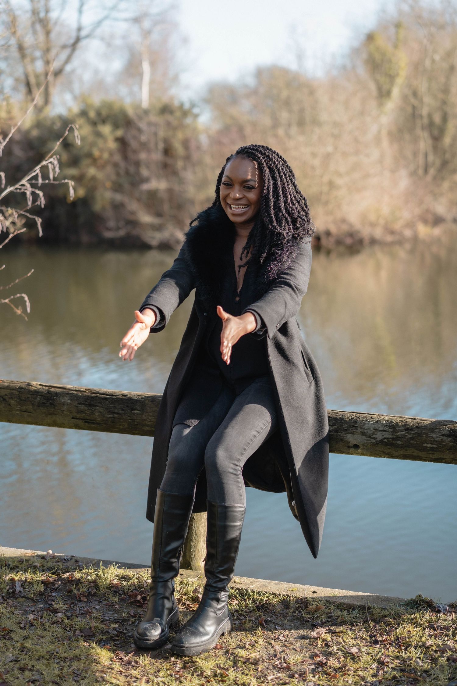 Person in a long grey coat poses expressively by a serene lake on an autumn day.
Mère tendant les bras et assise au bord d'un étant.