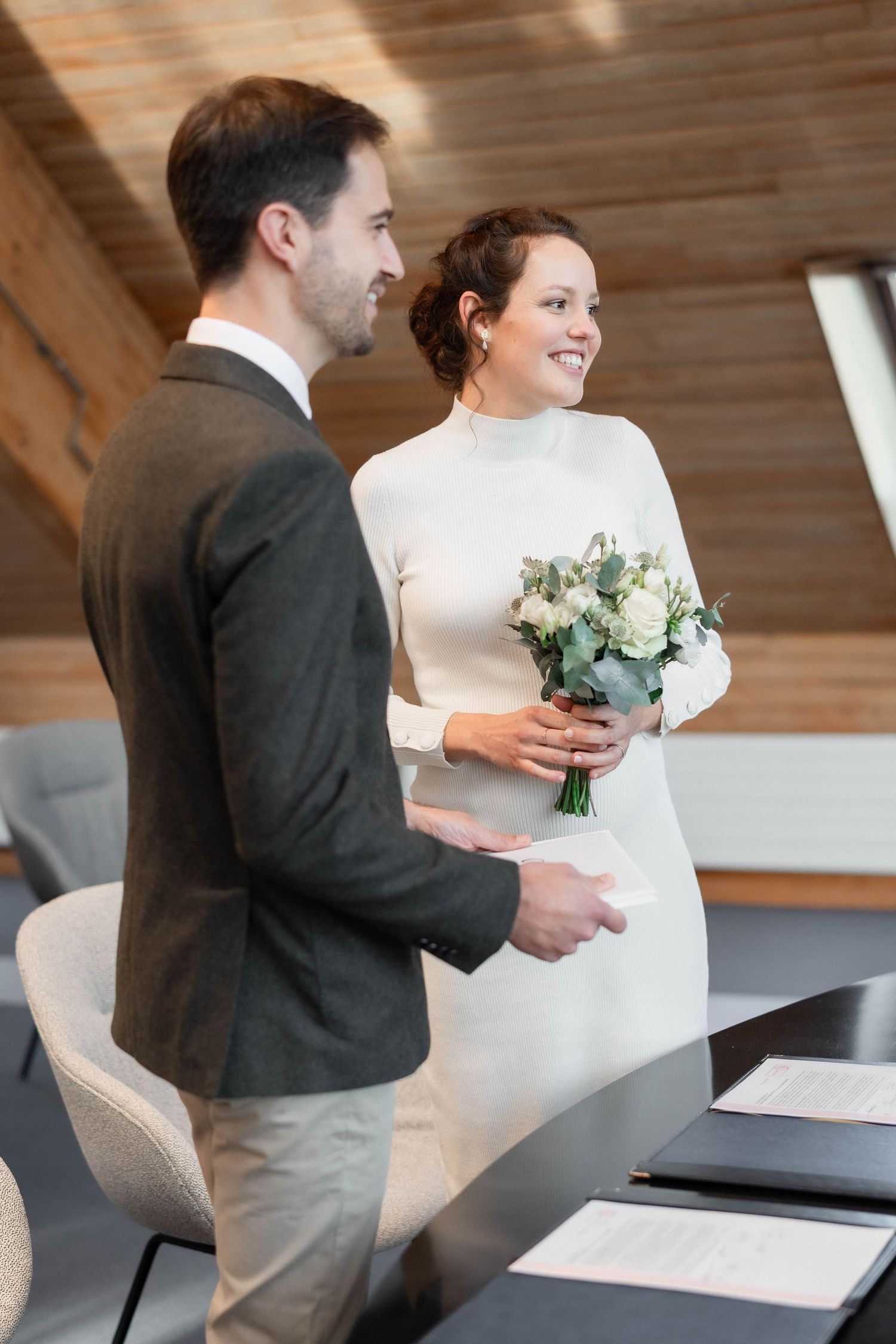 A couple smiling while signing papers in an office with wooden ceiling.
Mariés debout durant la cérémonie civile.