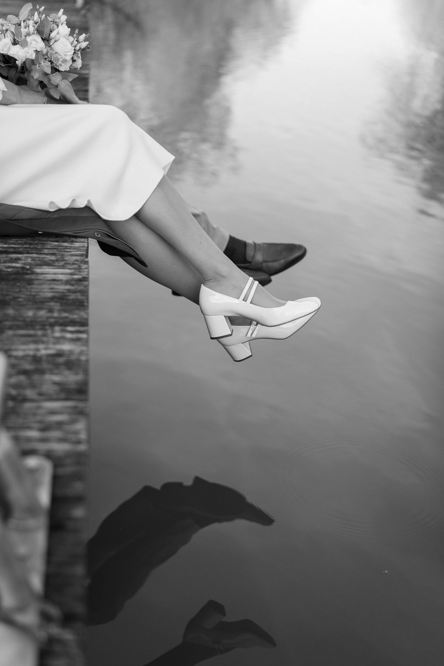 Artistic black and white photo of white bridal shoes dangling over reflective water.
Reflet jambes des mariés dans l'eau.