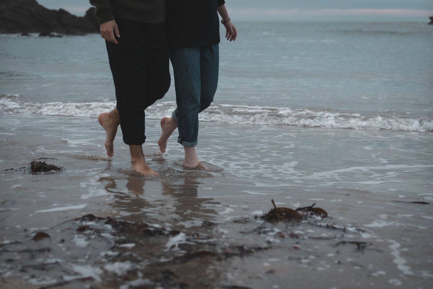 Two pairs of bare feet walking together in shallow ocean waves on a rocky beach.
Couple marchant dans l'eau.