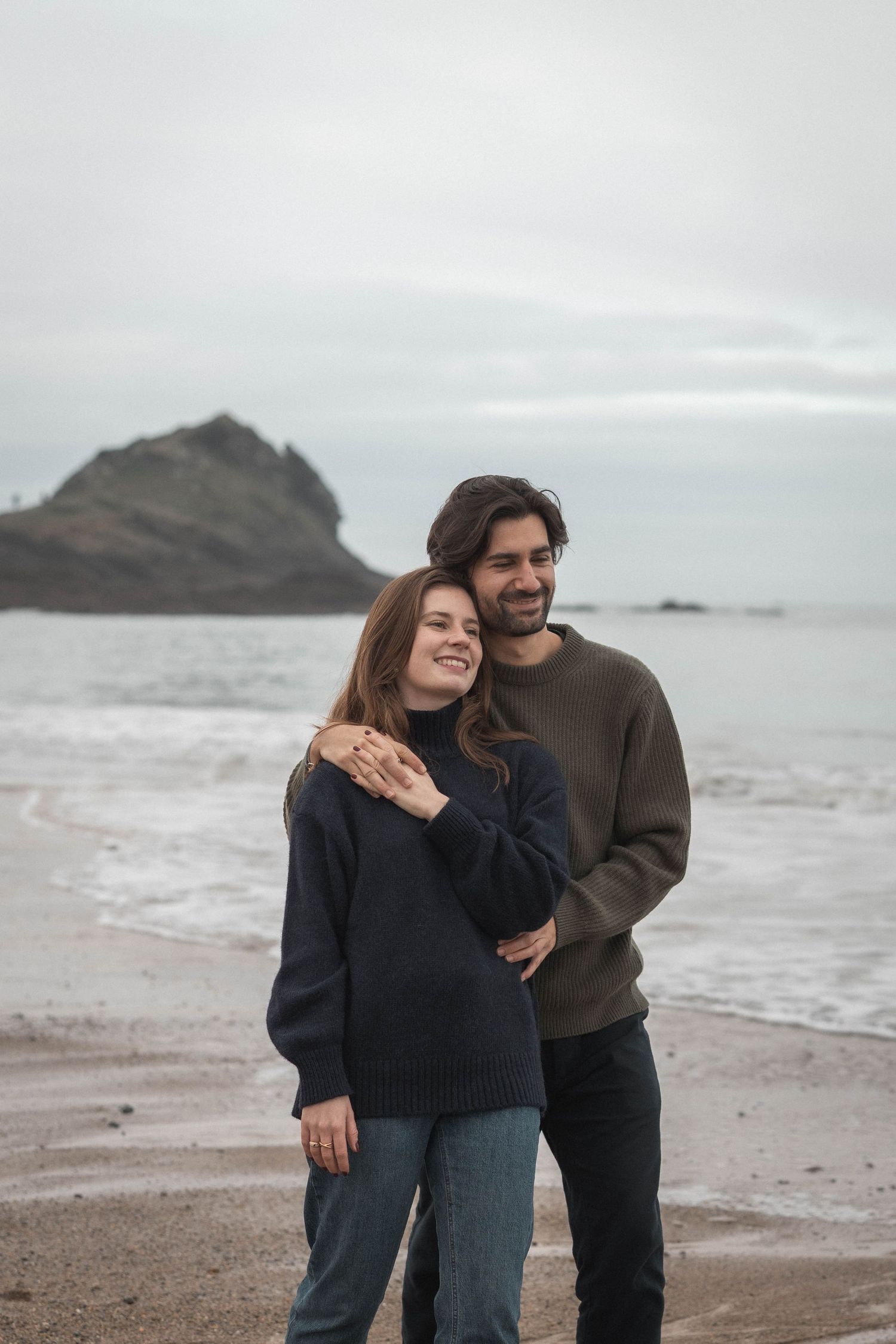 A couple shares a tender moment while standing on a cloudy beach with rocky cliffs in the background.
Couple sur la plage.