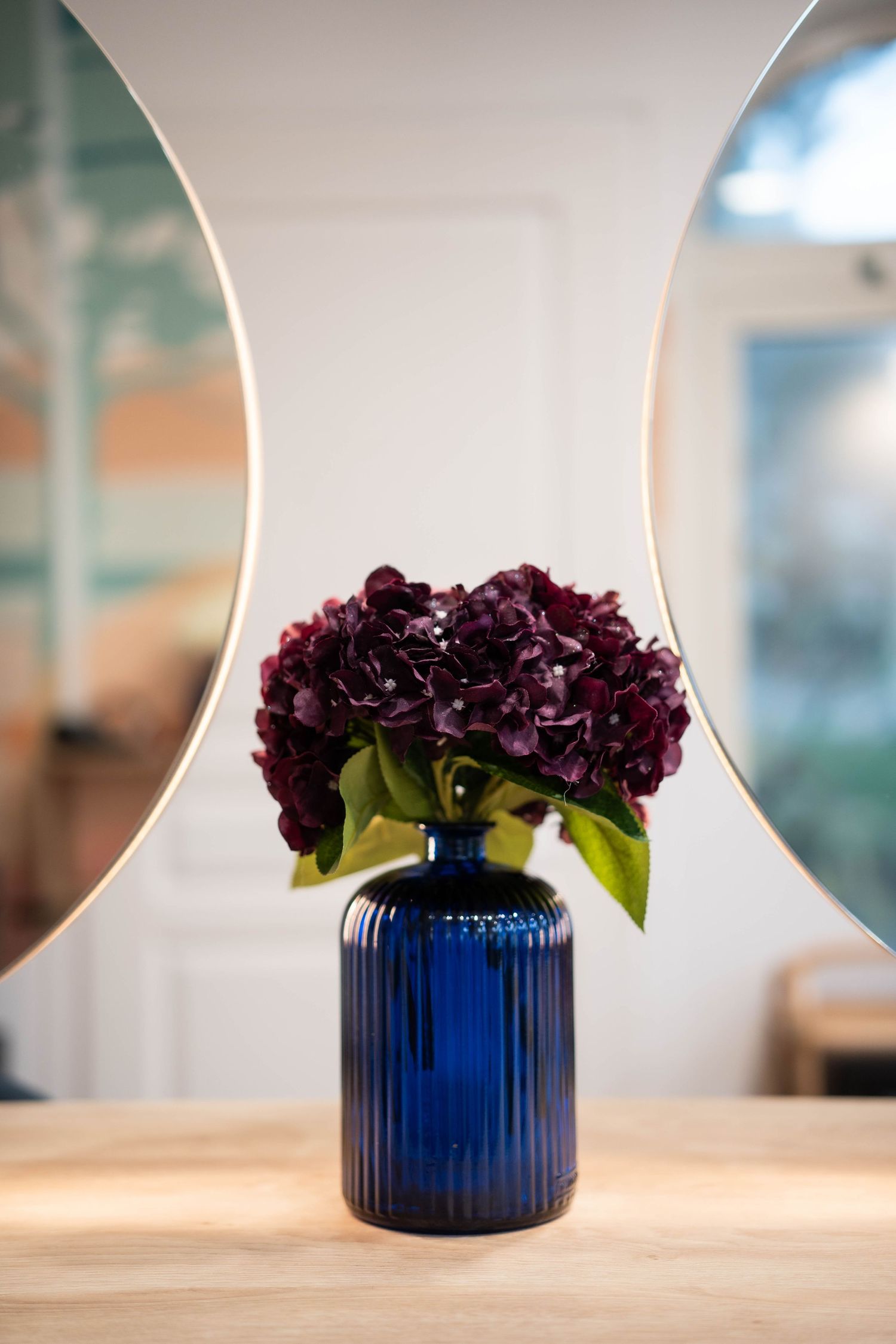 Deep purple hydrangeas in a ribbed blue glass vase on wooden surface.
Fleurs dans un vase posé entre deux miroirs.