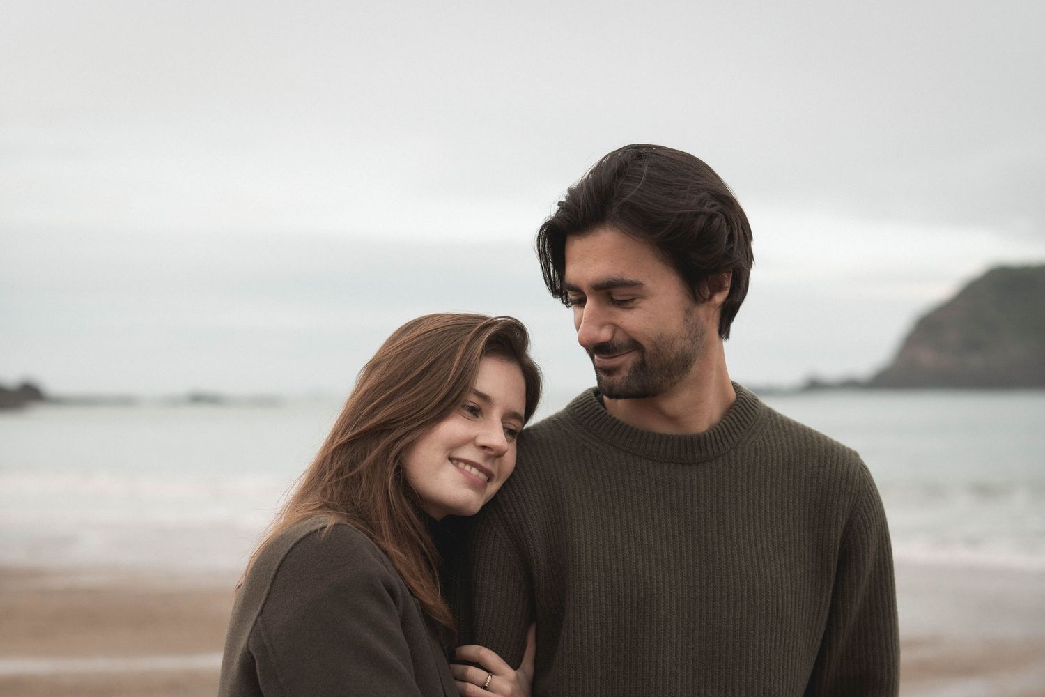 A couple in cozy sweaters shares a romantic moment on an overcast beach.
Couple au bord de la mer.