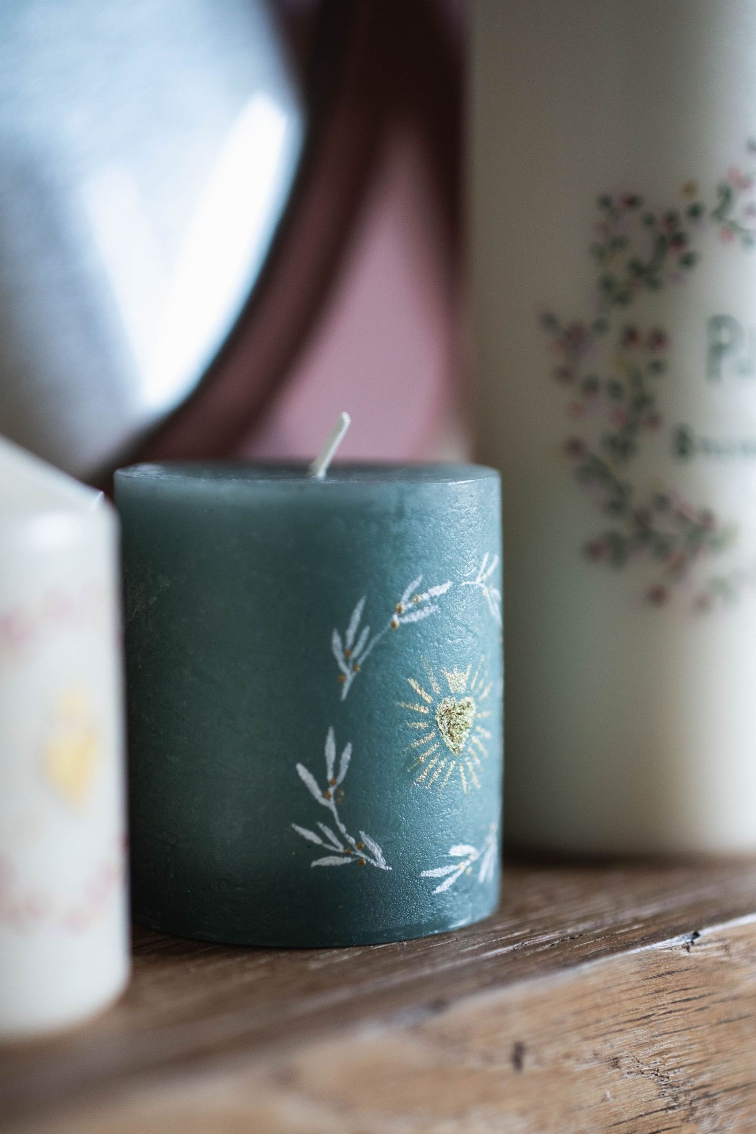 Close-up of a sage green candle with delicate floral and sun design etched in white and gold.
Bougies avec peintures florales.