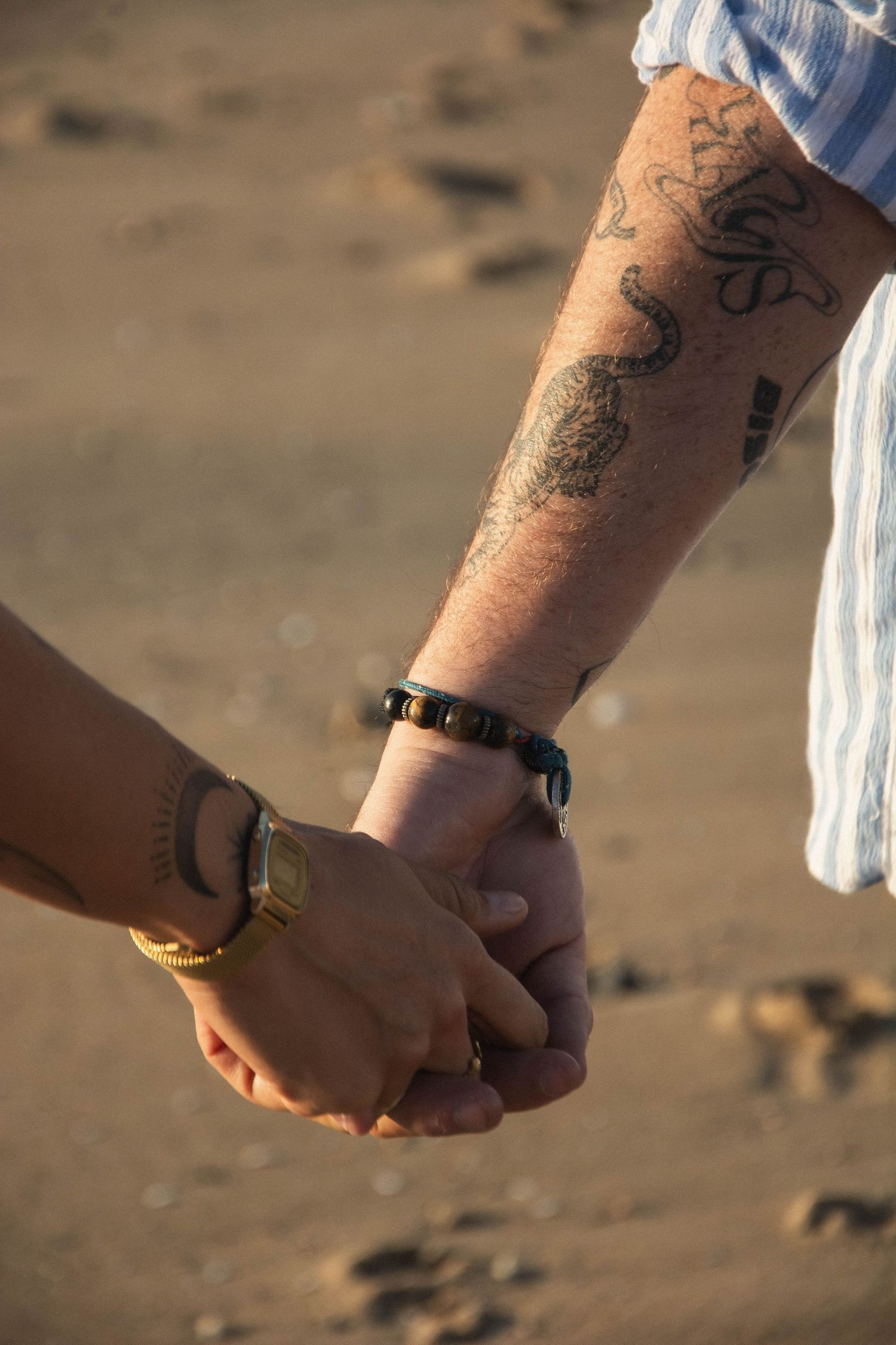 Close-up of two people holding hands with one person showing tattooed arm and bracelet on beach.
Mains se tenant à la plage, bras tatoués.
