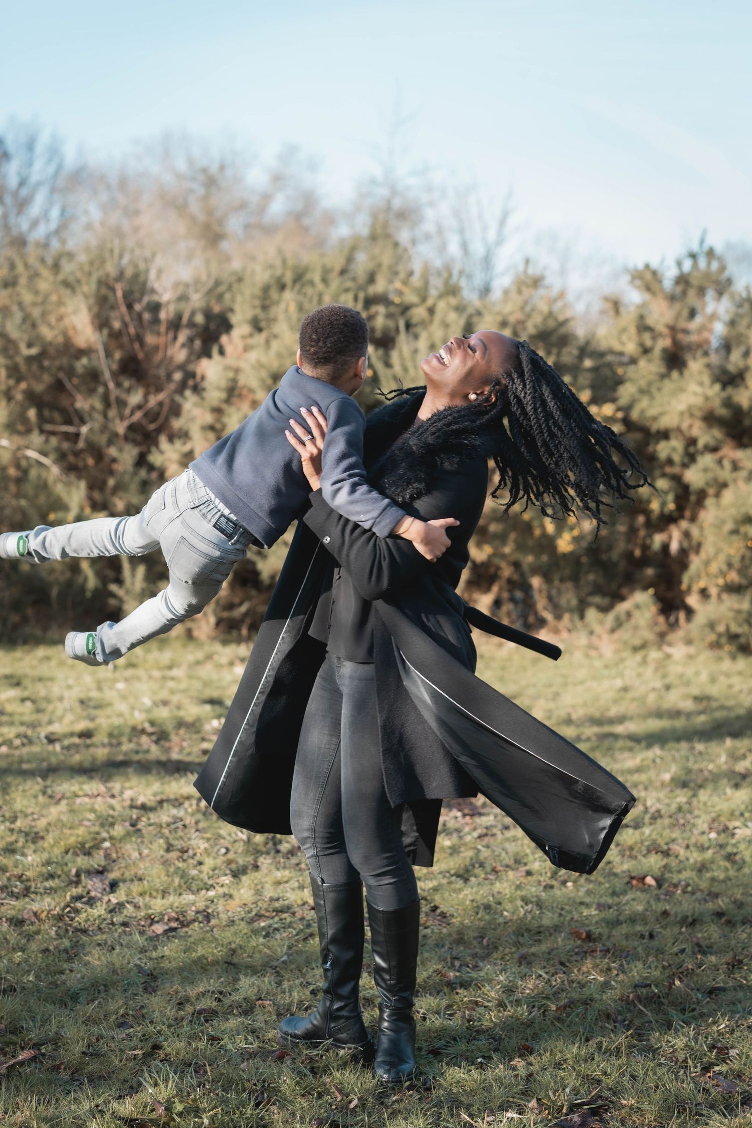 Two people in dark winter coats playfully spinning and dancing together in a grassy outdoor setting.
Mère tournant avec son fils dehors
