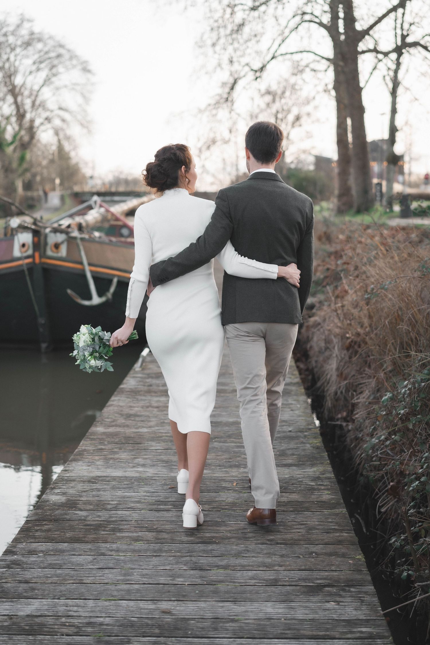 A newlywed couple walking arm in arm on a wooden dock beside a canal.
Couple de mariés de dos marchant au bord d'un canal.