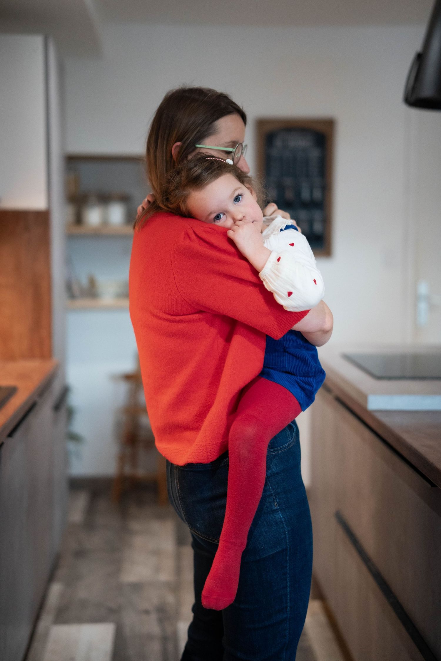 Mother in red sweater and blue jeans holding daughter while standing in a room.
Câlin d'une maman et sa fille debout dans la cuisine