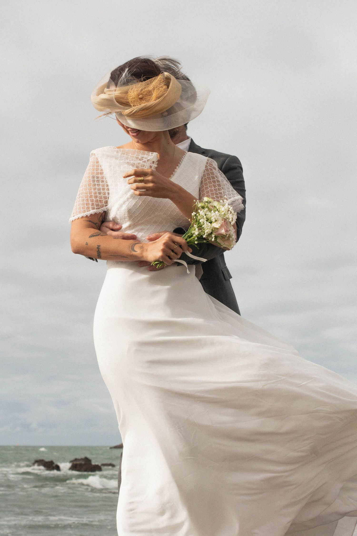 Romantic beach wedding scene with flowing white dress and hat caught in ocean breeze.
Couple mariés à la plage s'enlaçant devant l'océan.
