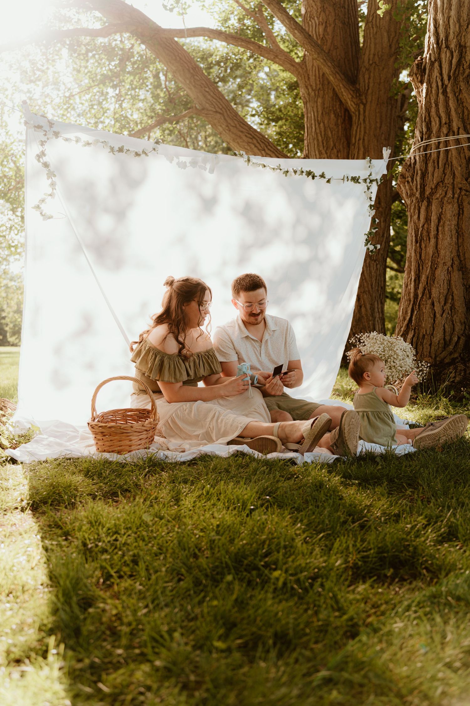 Family enjoying a sunny outdoor picnic on grass under large trees with a white backdrop.