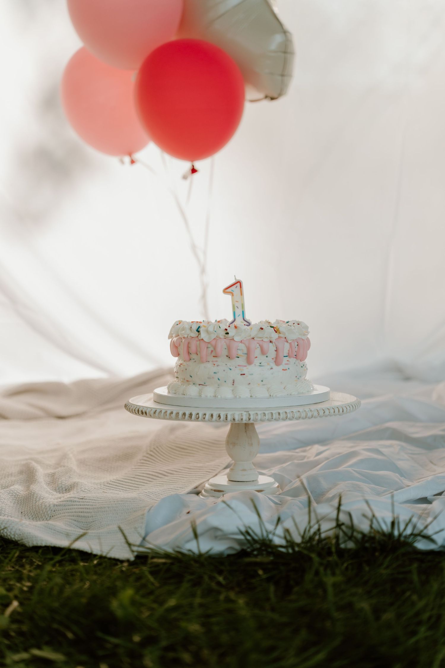 A white first birthday cake with a number 1 candle on a cake stand, surrounded by pink and red balloons outdoors.