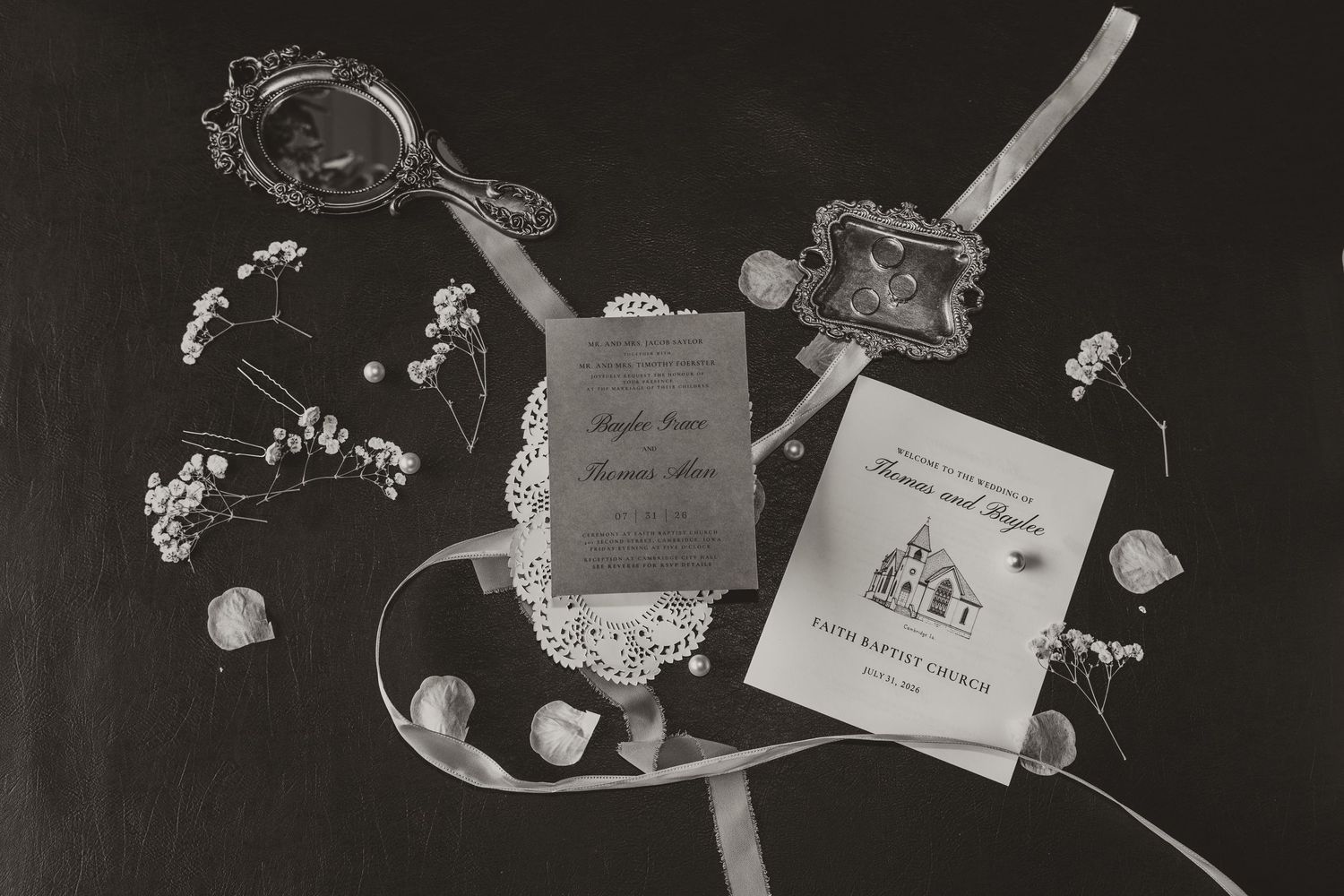Black and white wedding flat lay with invitations, hand mirror, ribbon, and floral accents on dark background.