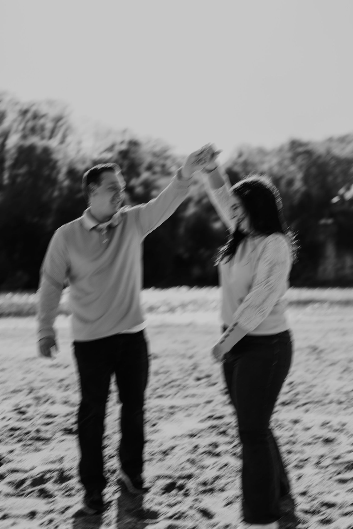 Black and white photo of a couple dancing outdoors, man spinning woman with raised hands in a field with trees behind.