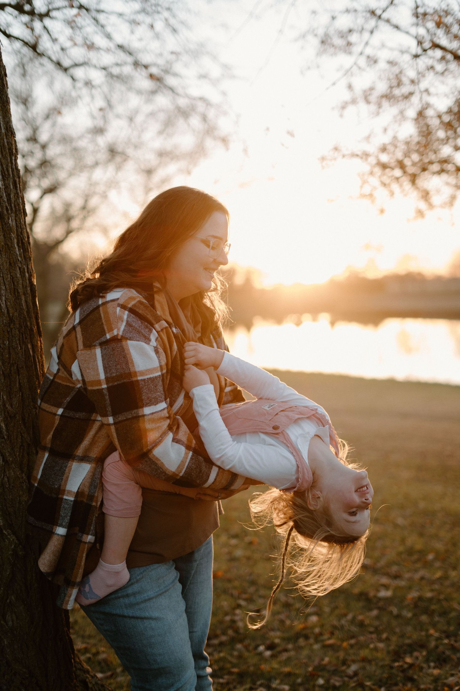 Mother playfully holding child upside down near tree at golden sunset by a lake in autumn park.