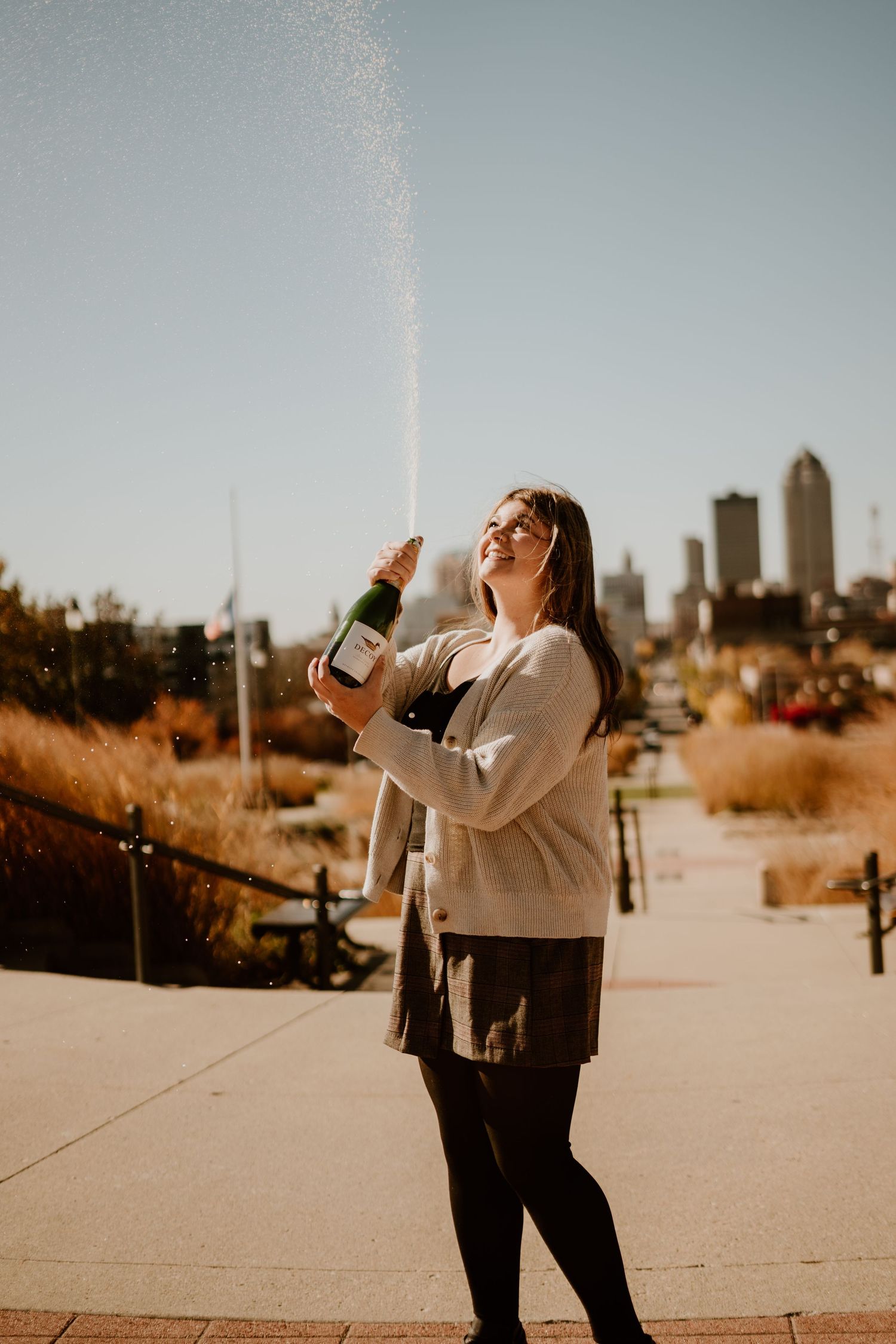 Woman joyfully popping champagne bottle outdoors with city skyline in background on a sunny day.