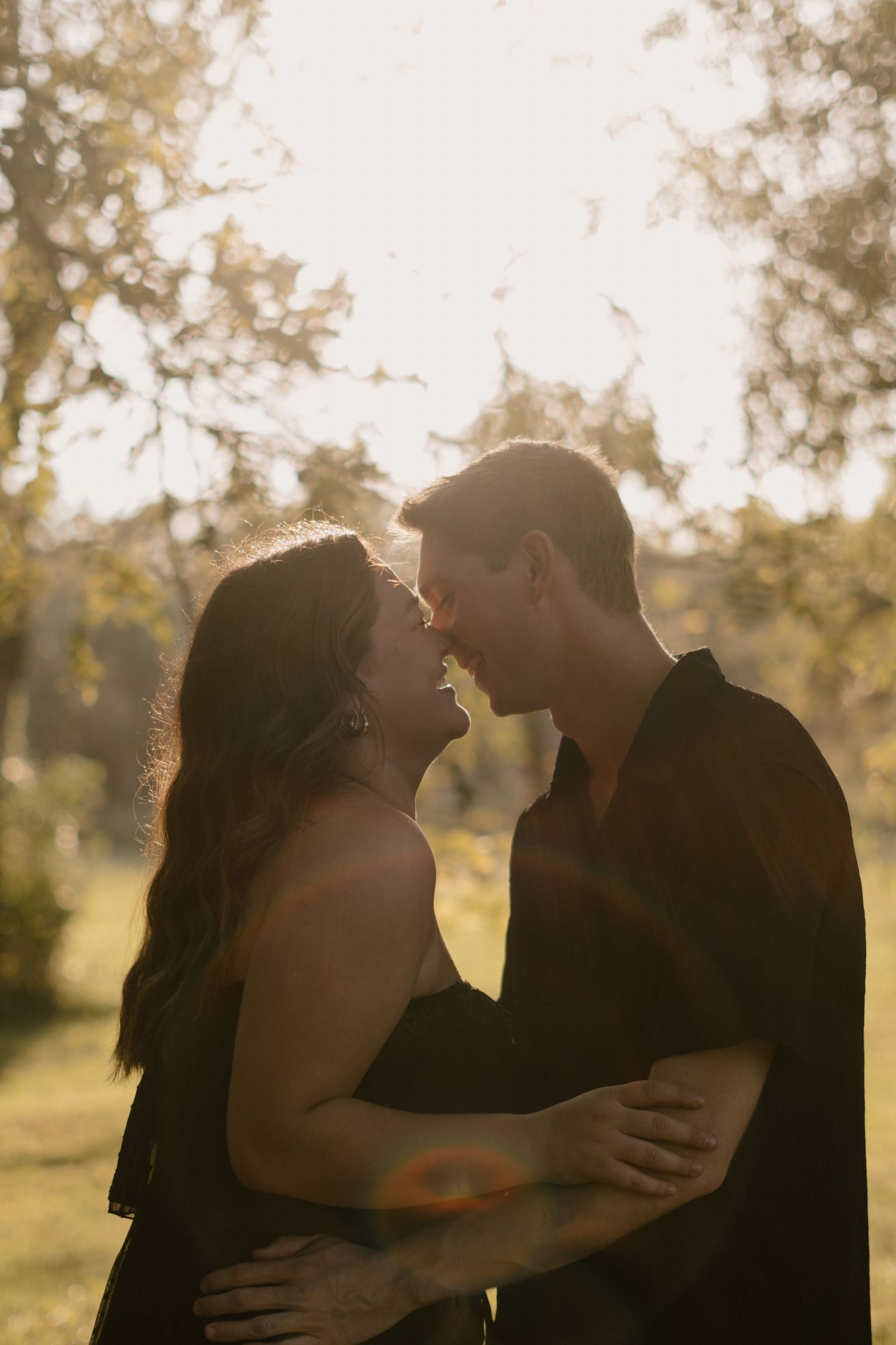 A couple in black embraces and leans in for a kiss, bathed in warm golden sunlight in a lush green park.